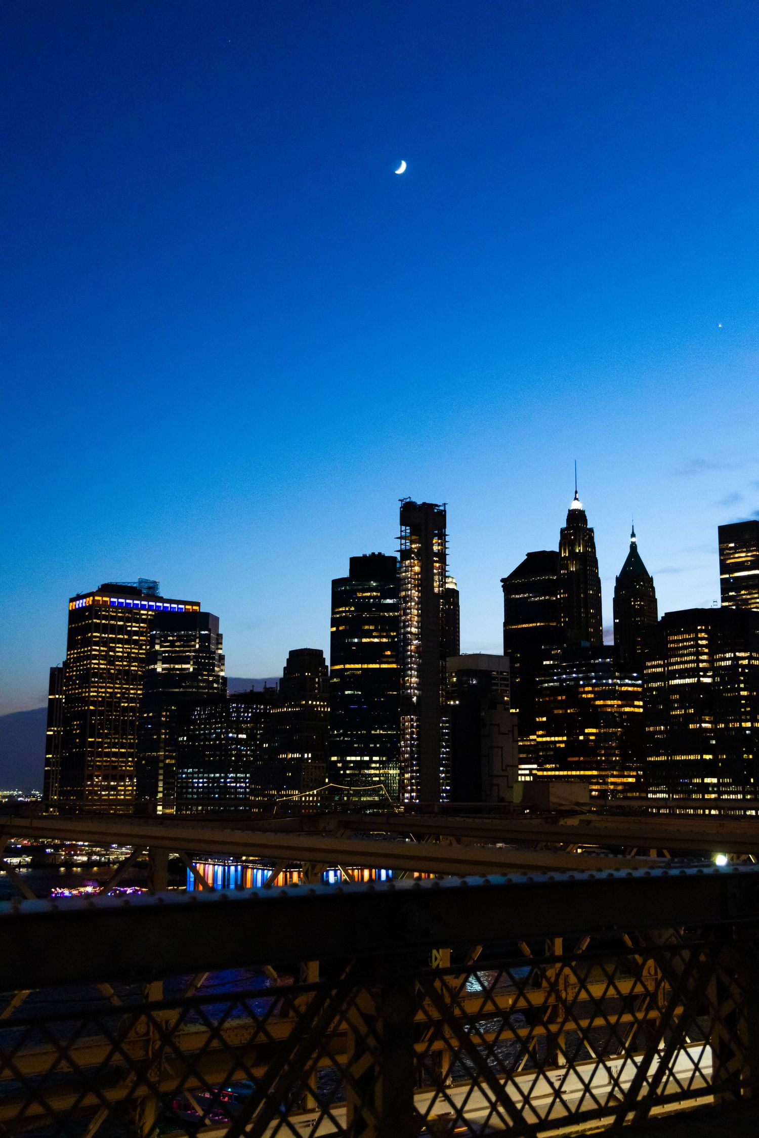 NYC skyline glowing at dusk with crescent moon, viewed from Brooklyn Bridge, skyscrapers illuminated against deep blue sky.