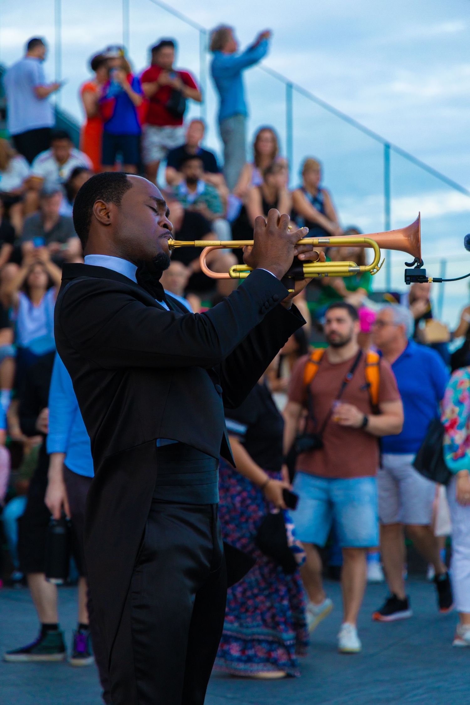 A Black musician in a black suit plays a gold trumpet outdoors for a large crowd at a live music event.