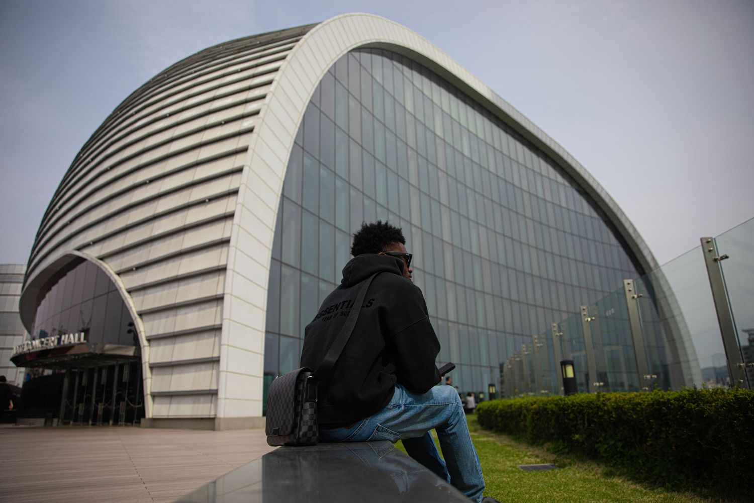 Person sitting outside a modern curved glass building with an arched facade in an urban setting.