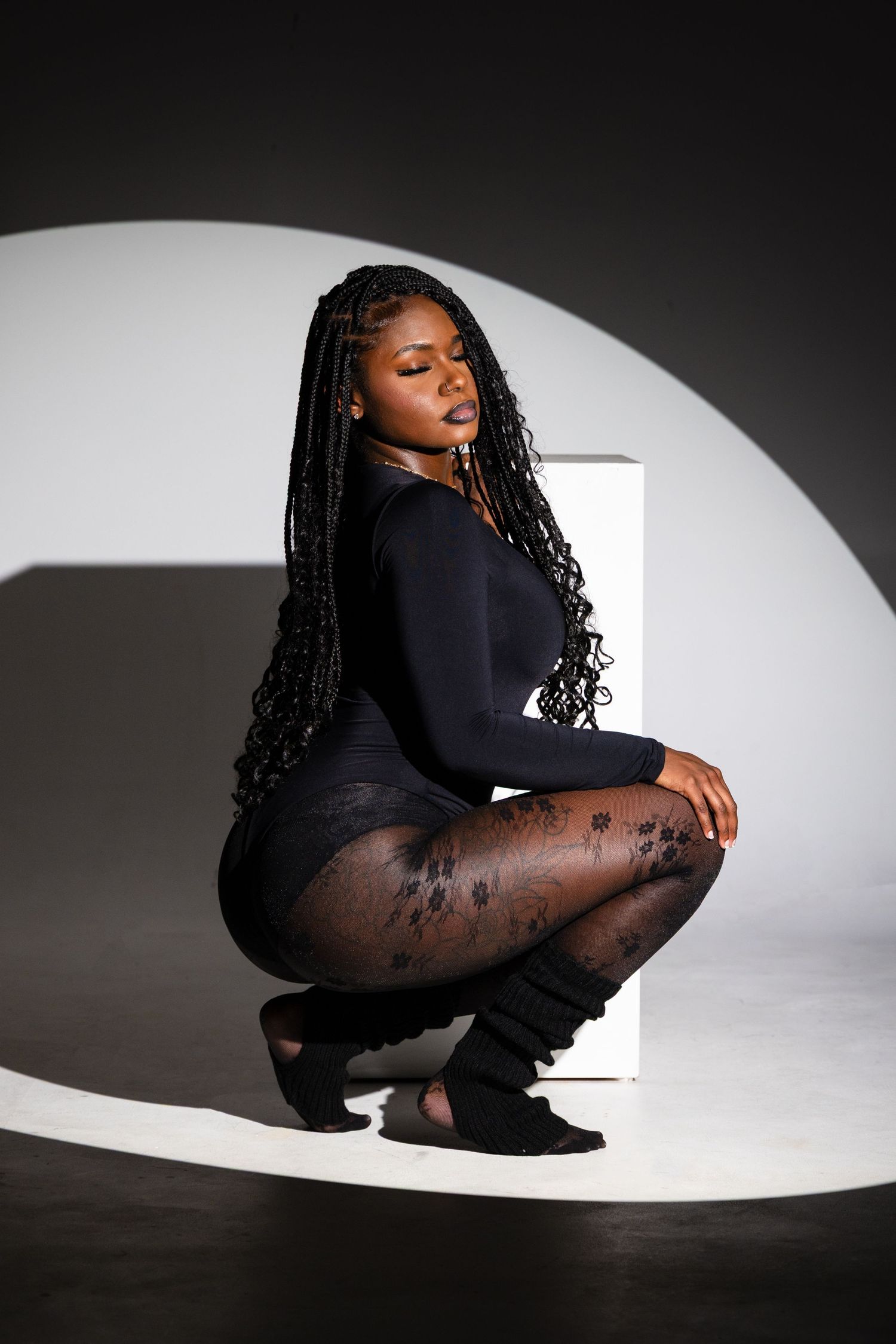 A woman with long box braids crouches in a black bodysuit and floral tights against a studio backdrop.