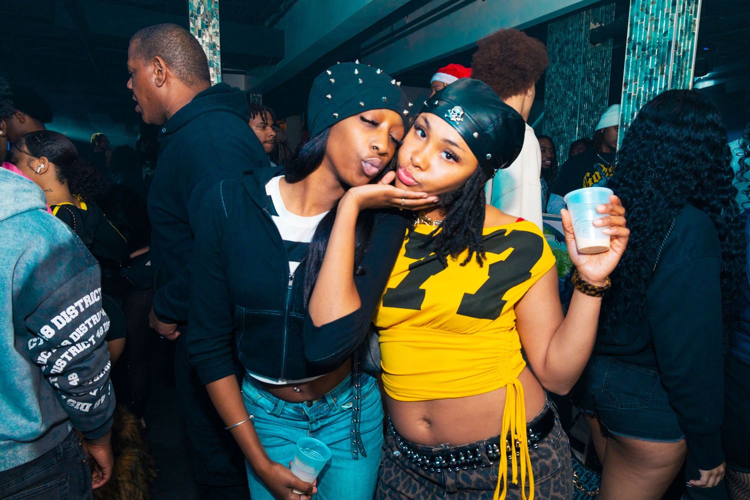 Two young women pose together at a lively nightclub party, one wearing a yellow crop top holding a drink.