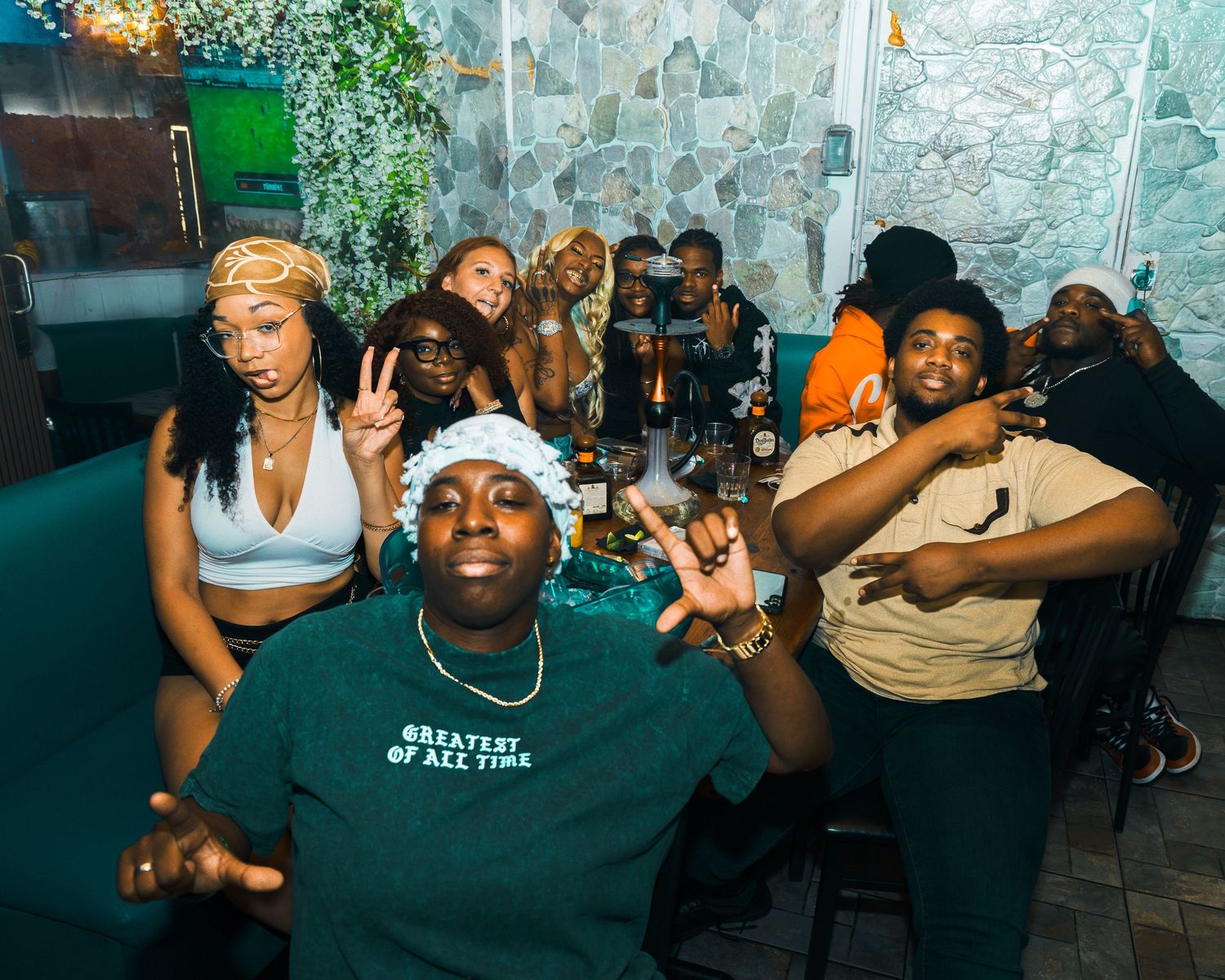 Group of friends posing and smiling at a restaurant booth, one wearing a Greatest of All Time green shirt.