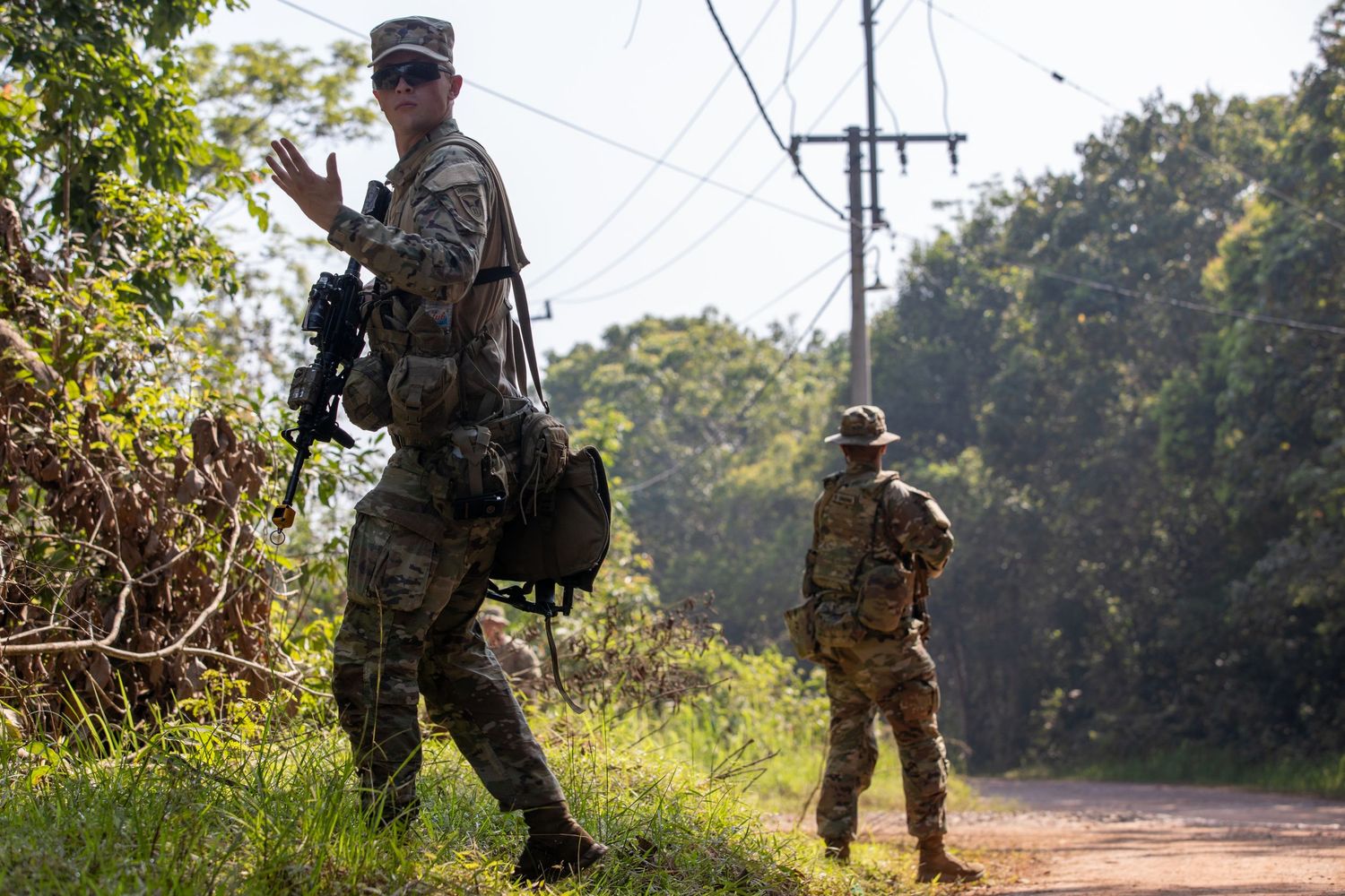 Armed US military soldiers in tactical gear signal during a patrol exercise along a rural jungle road.
