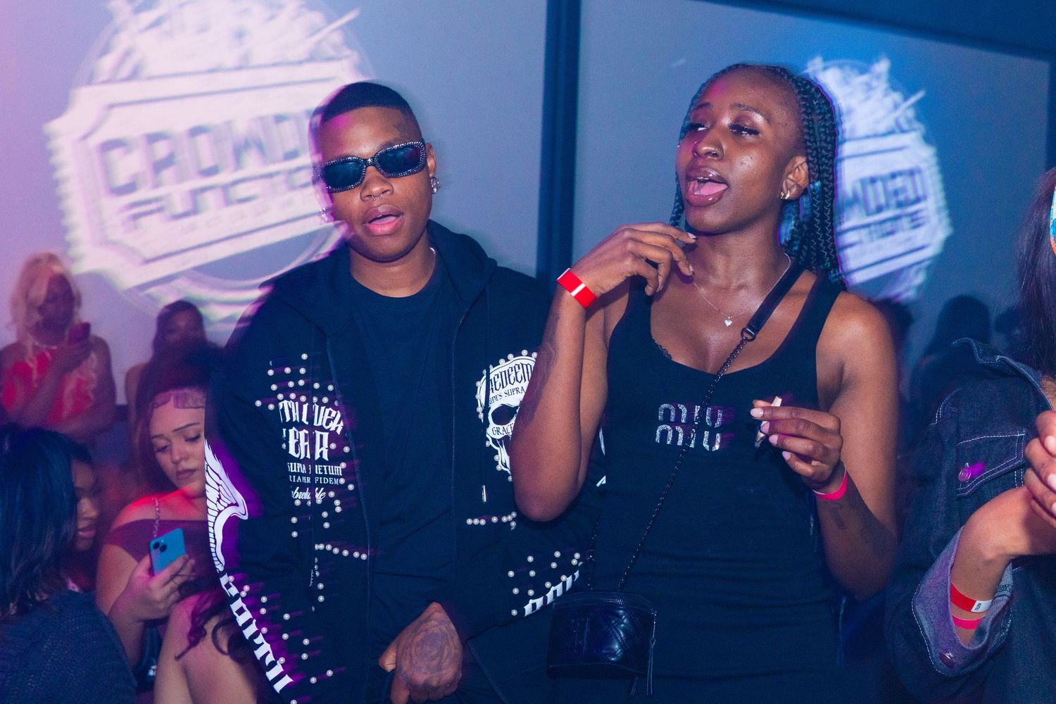 Two people posing at a Crowd Funded event, wearing stylish black outfits in a vibrant nightclub setting.