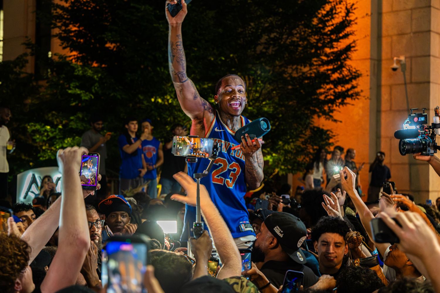 Crowd celebrating at night with man in blue jersey #23 raised on shoulders, fans cheering with phones out.