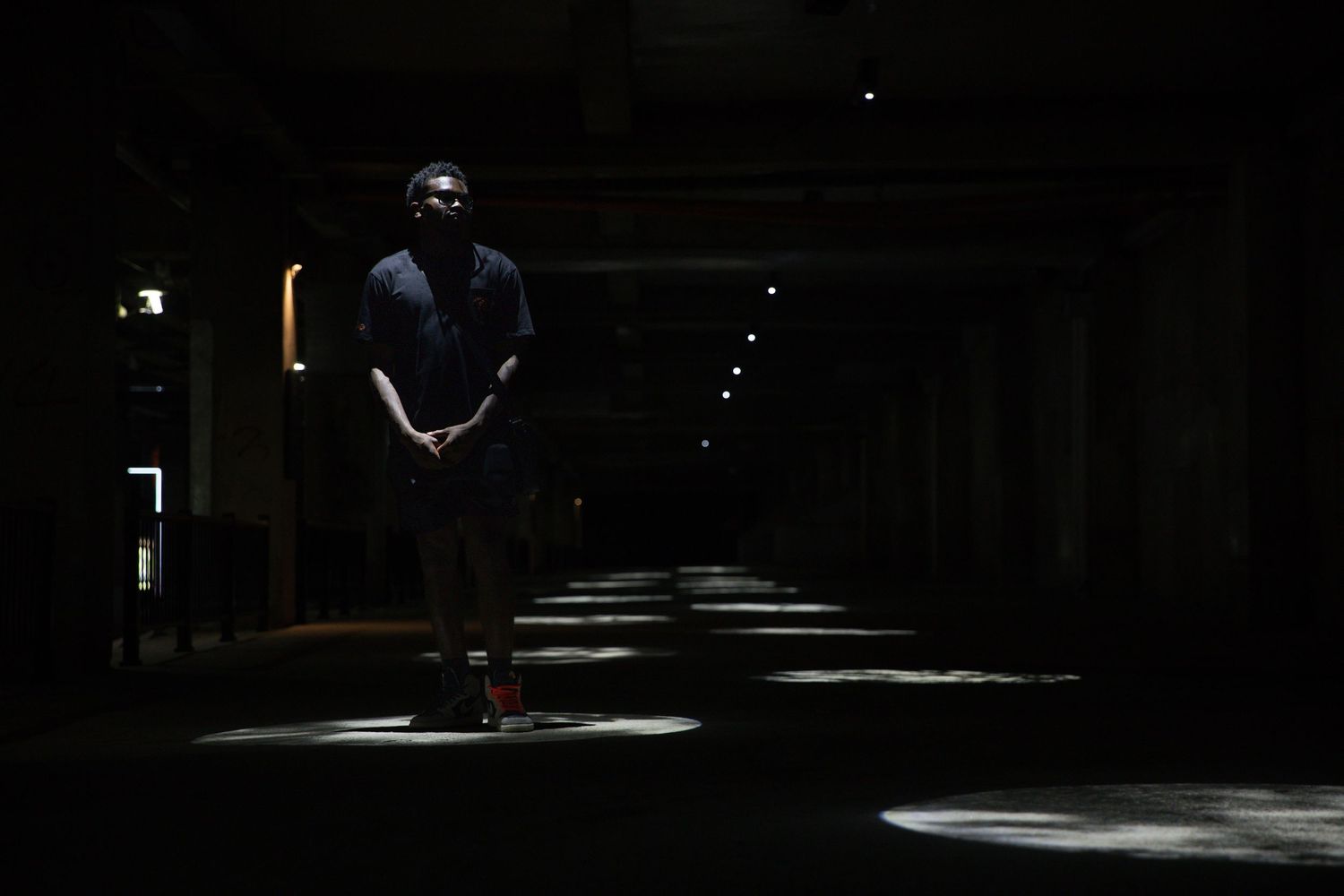 A lone figure stands illuminated by a single spotlight in a dark, atmospheric corridor with scattered light pools.