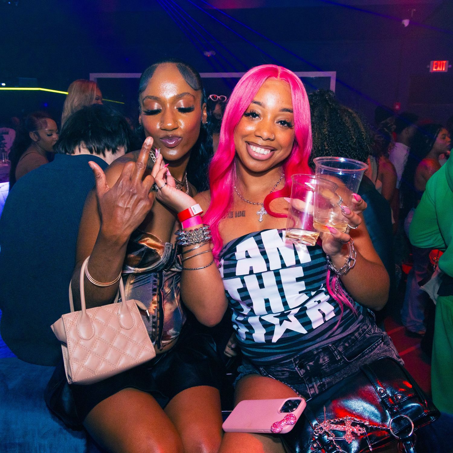 Two women posing at a nightclub, one with pink hair holding a cup, wearing colorful outfits and accessories.