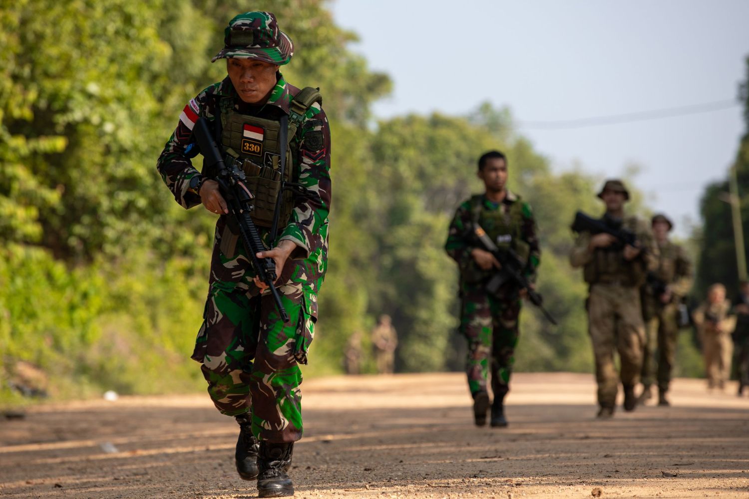 Indonesian soldiers in camouflage uniforms patrol a dirt road during a military training exercise in a tropical environment.