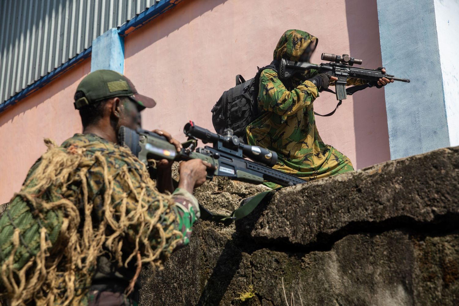Two camouflaged snipers aiming rifles from behind concrete barrier during military training exercise.