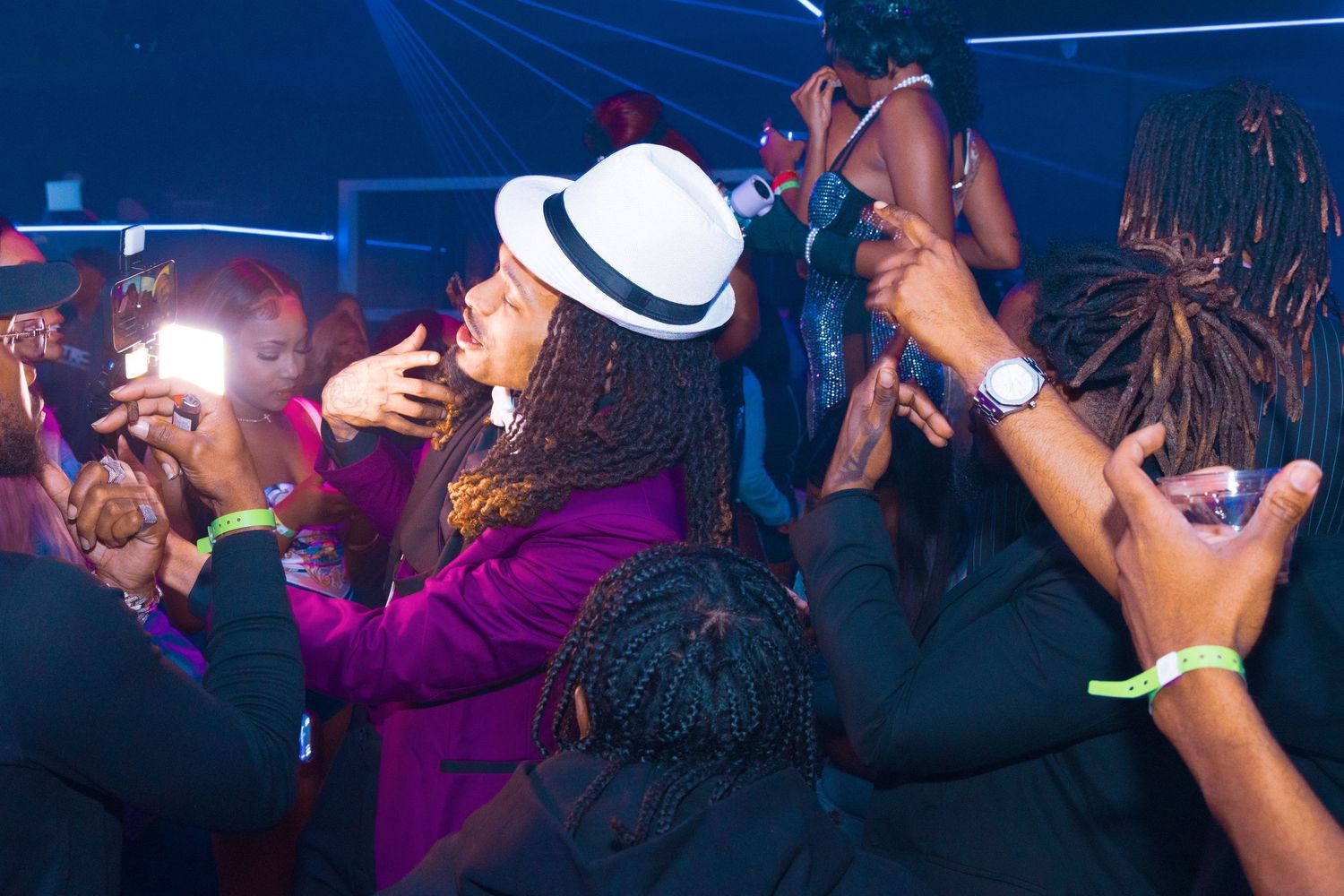 People dancing at a lively nightclub party, with colorful lights and a woman in a white hat and purple jacket.