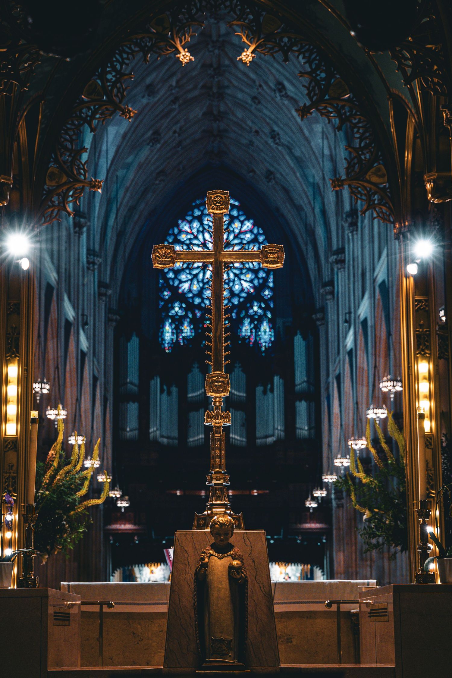 Golden cross altar in gothic cathedral with blue rose window and organ pipes illuminated by candlelight.