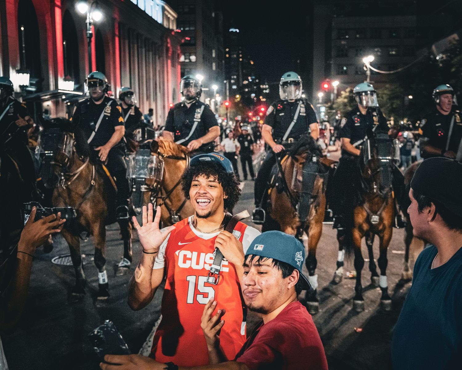 Two young men smile and pose in front of mounted police officers on horseback at a nighttime urban event.
