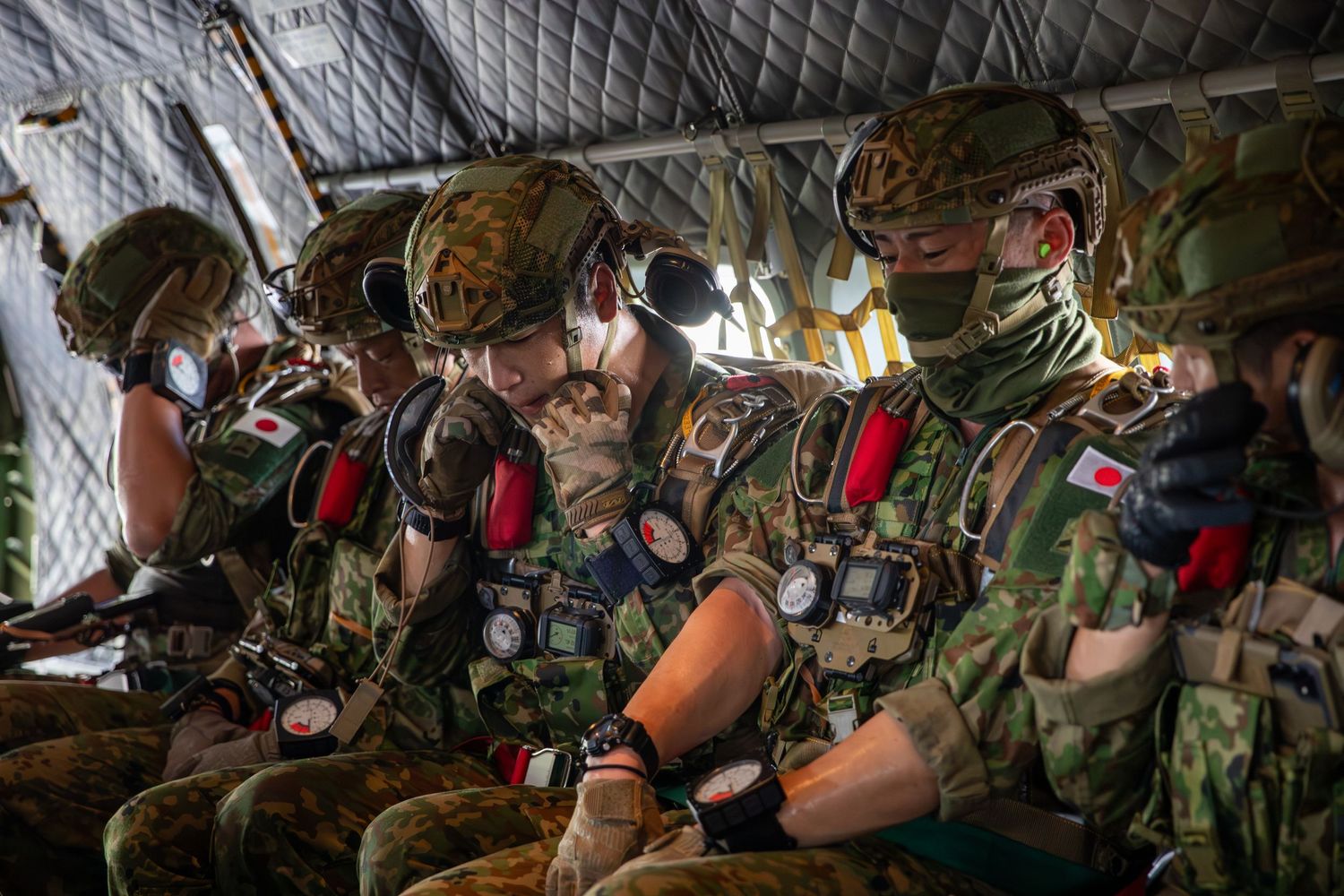 Japanese military paratroopers in camouflage gear prepare equipment inside an aircraft before a jump.