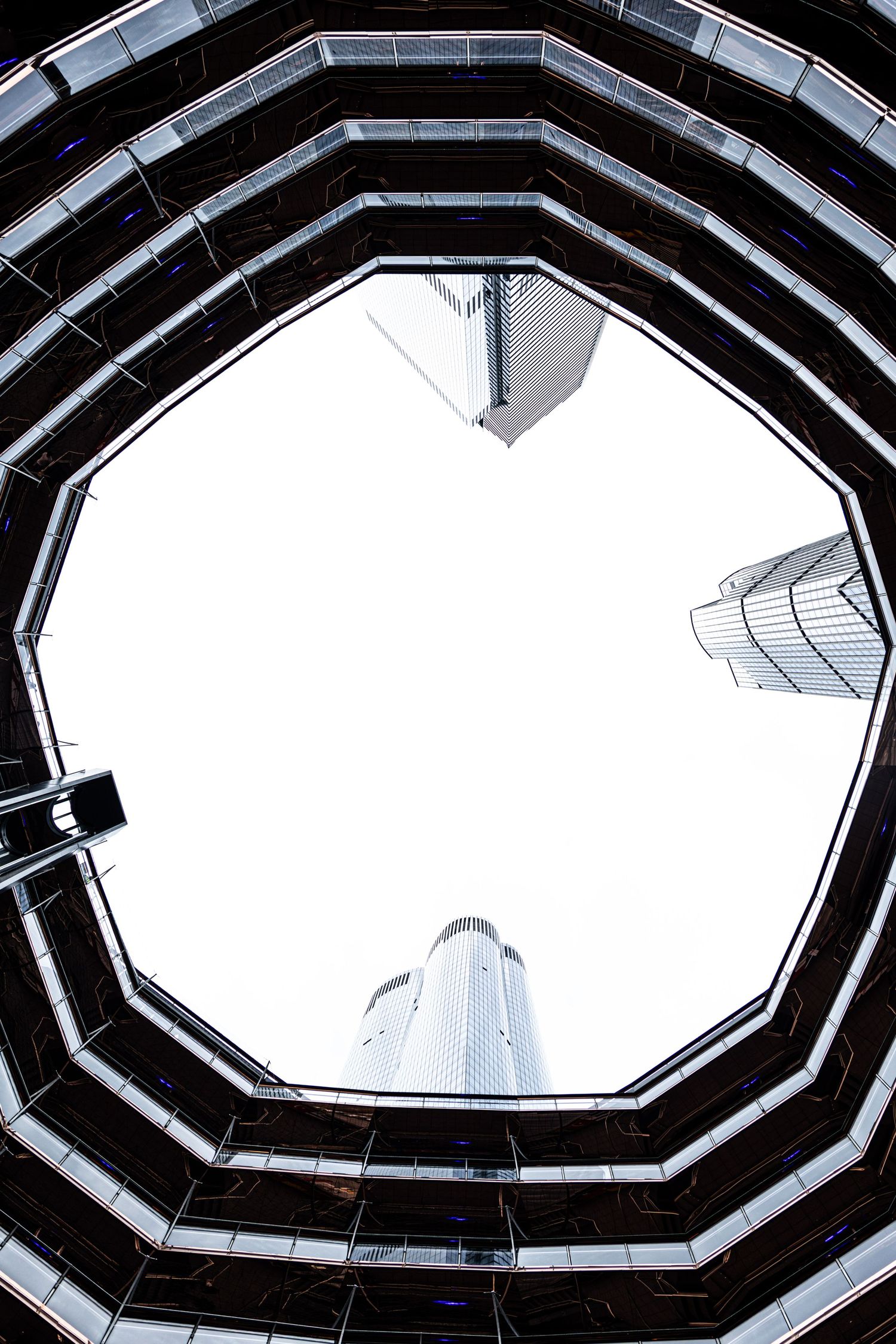 Looking up through an octagonal glass atrium at modern skyscrapers against a bright white sky.