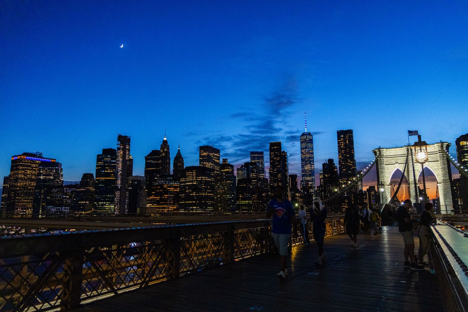 People on Brooklyn Bridge at dusk with NYC skyline illuminated against deep blue twilight sky.