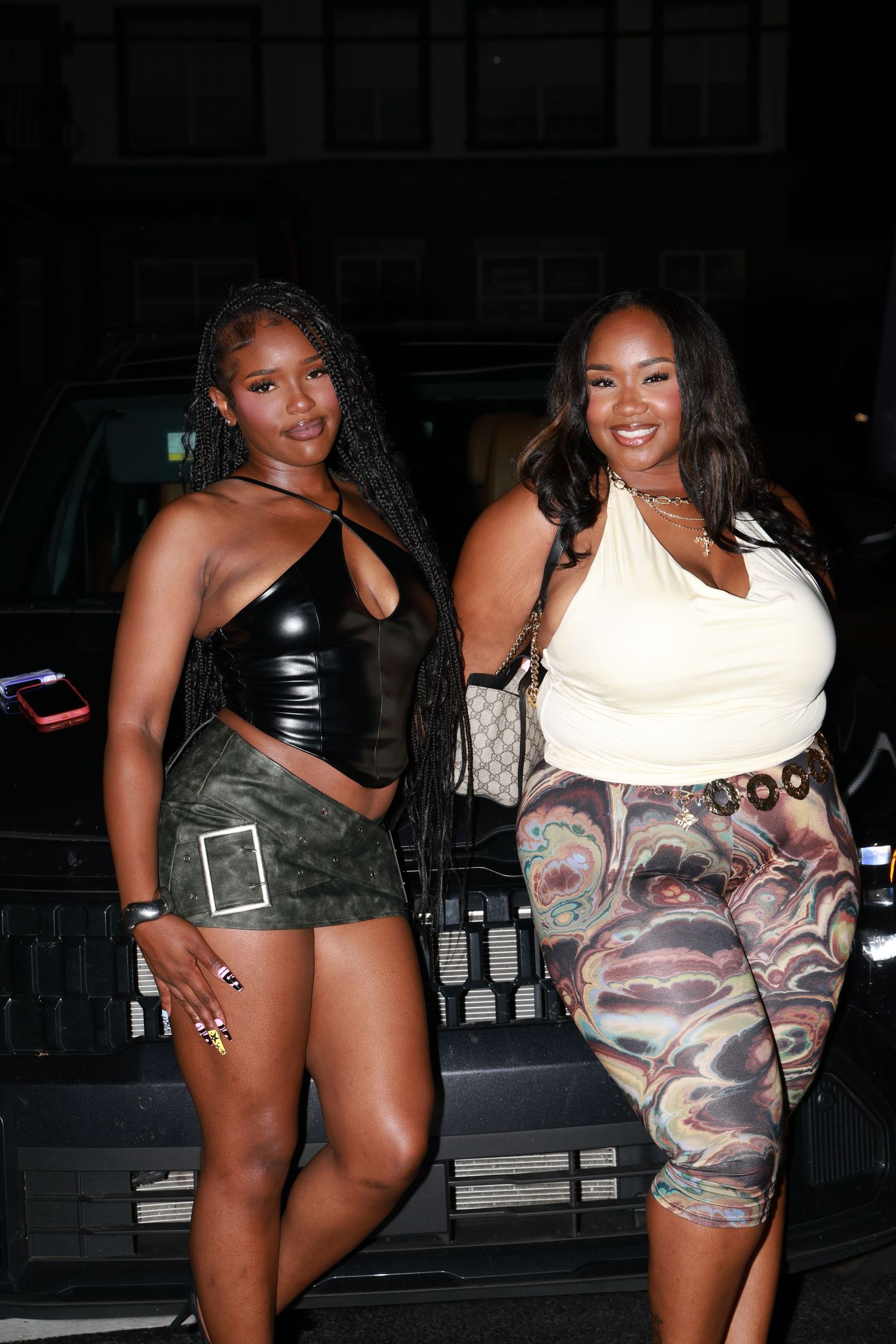 Two stylish women posing outdoors at night, one in black leather crop top with mini skirt, other in white top and printed leggings.
