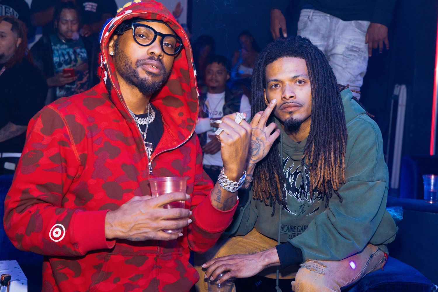 Two men posing together at a nightclub event, one in a red camo hoodie holding a drink.
