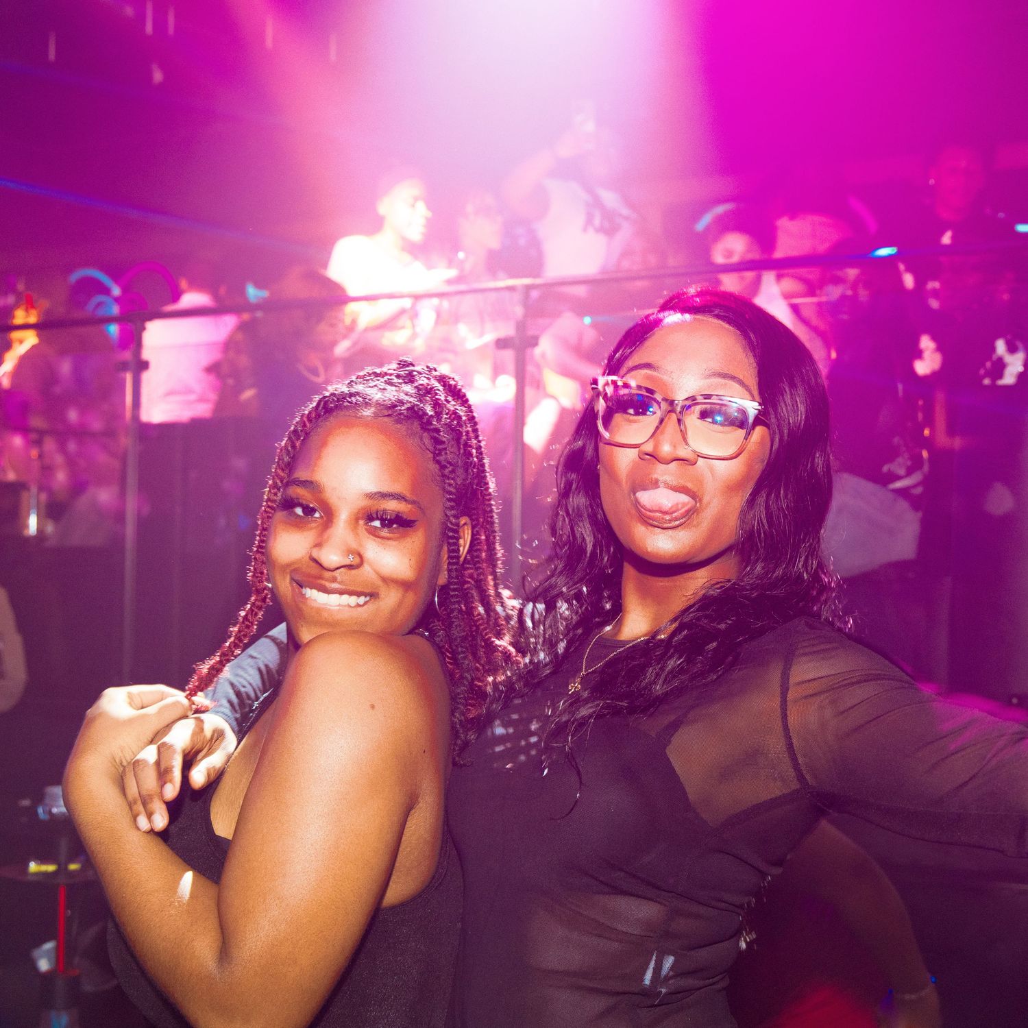 Two women smiling and posing together at a nightclub with colorful pink and orange lights in the background.
