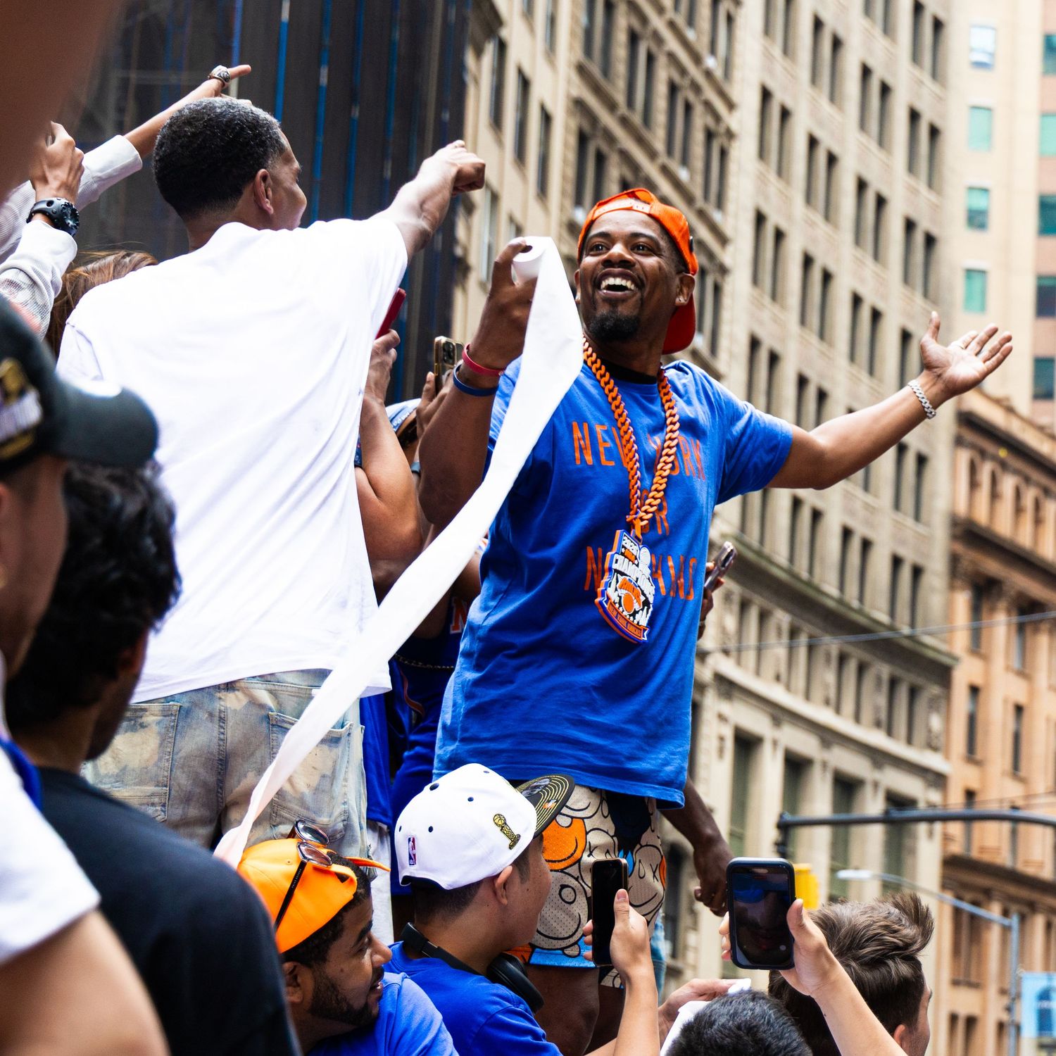 Man in blue shirt celebrating at a street parade, raising arms joyfully amid a crowd in an urban setting.
