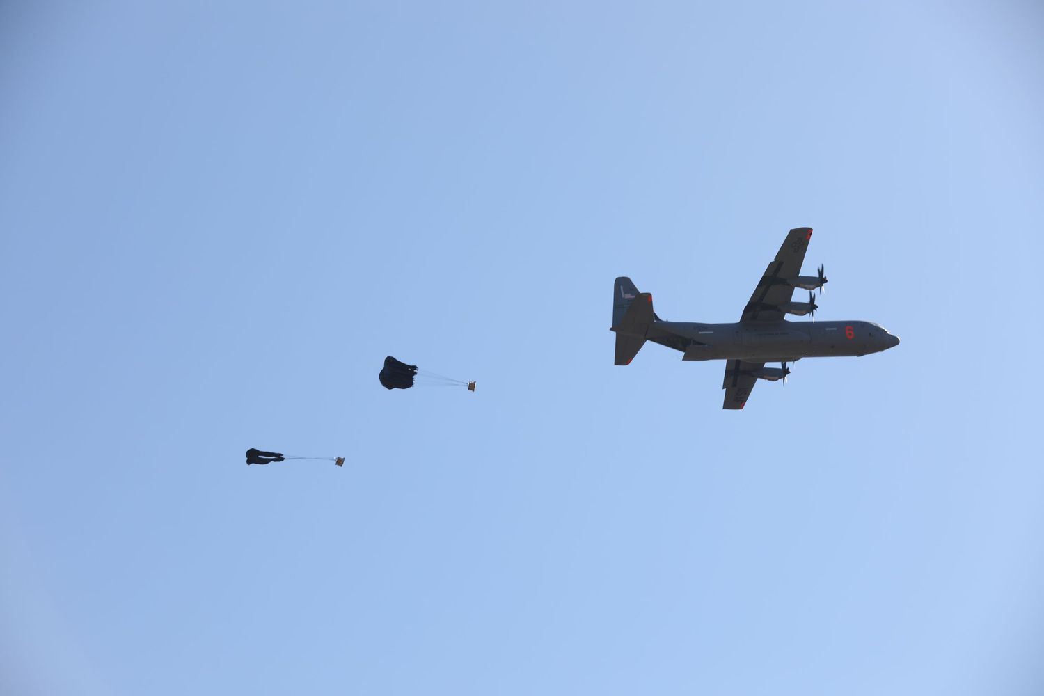A military C-130 aircraft drops cargo bundles over a clear blue sky during an airdrop training exercise.