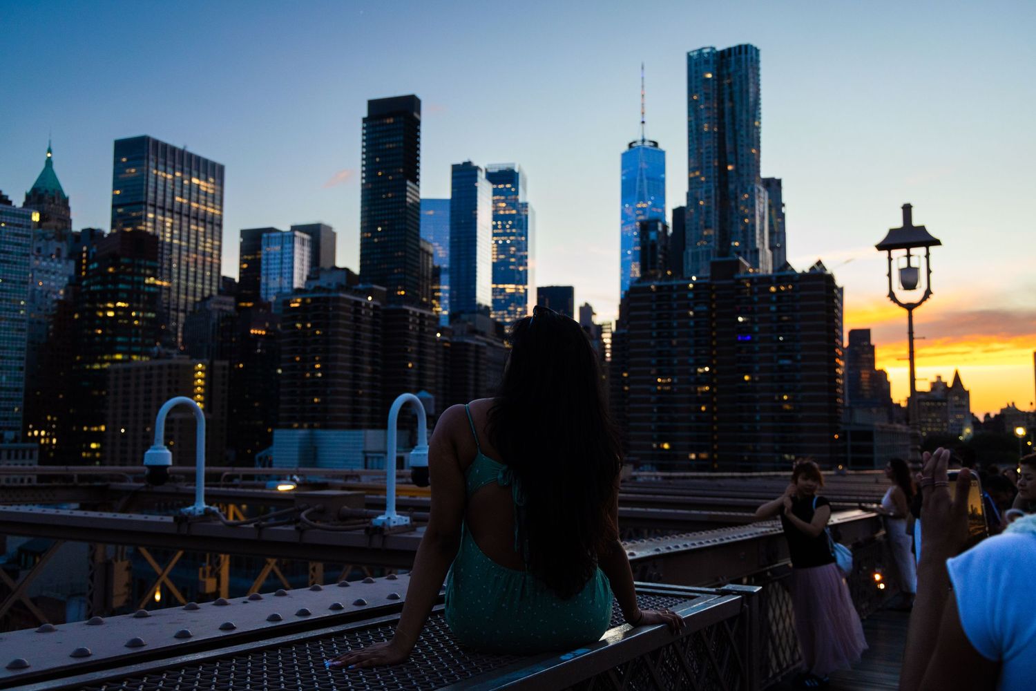 Woman admiring NYC skyline with One World Trade Center at sunset from Brooklyn Bridge.