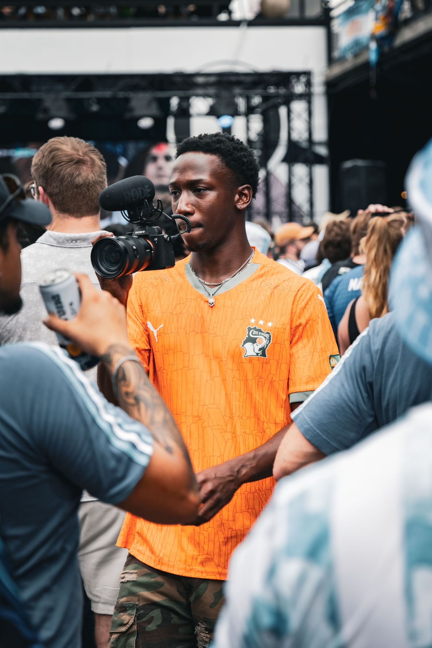 Young man in orange soccer jersey holds a camera at an outdoor music festival crowd.