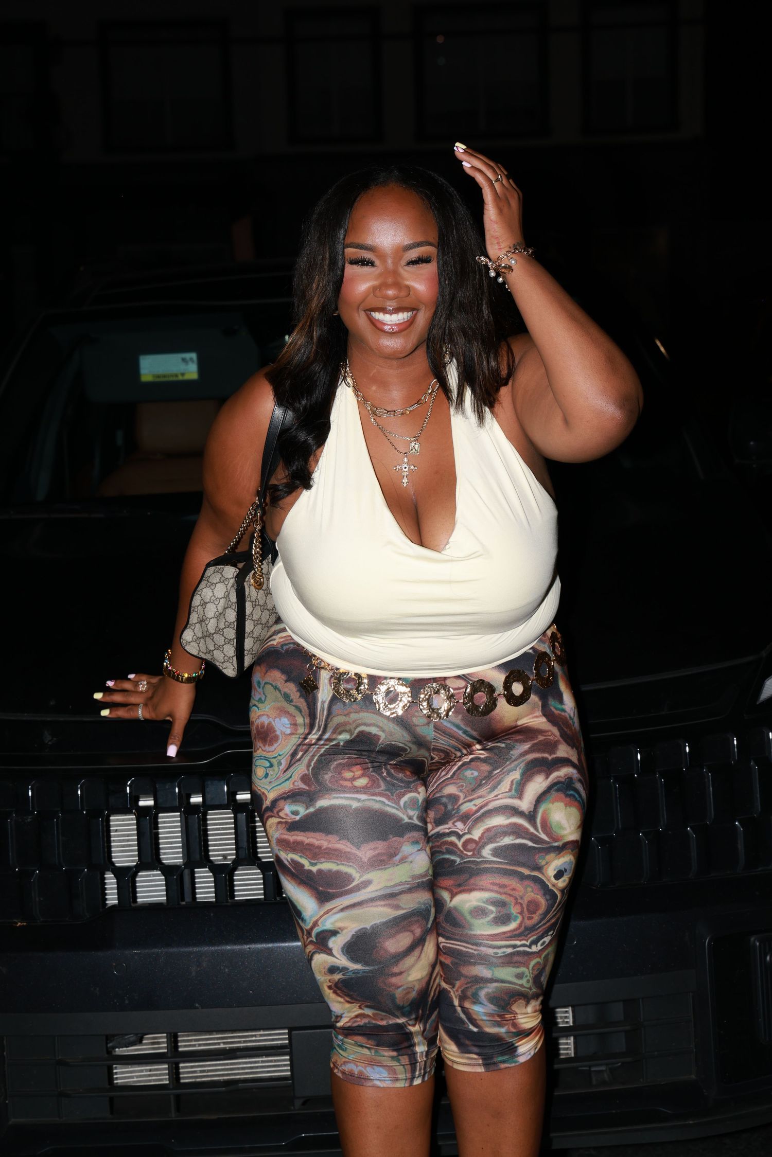 Smiling woman in white halter top and colorful printed biker shorts poses outdoors at night near a dark vehicle.