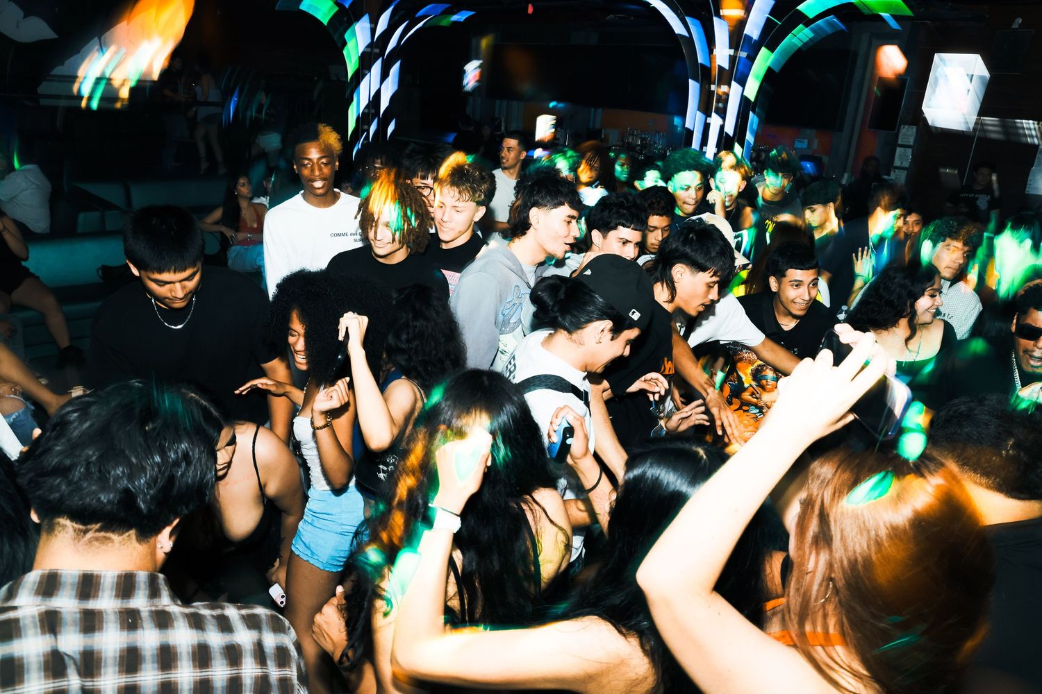 Crowded nightclub dance floor with young people dancing under green and blue lights.