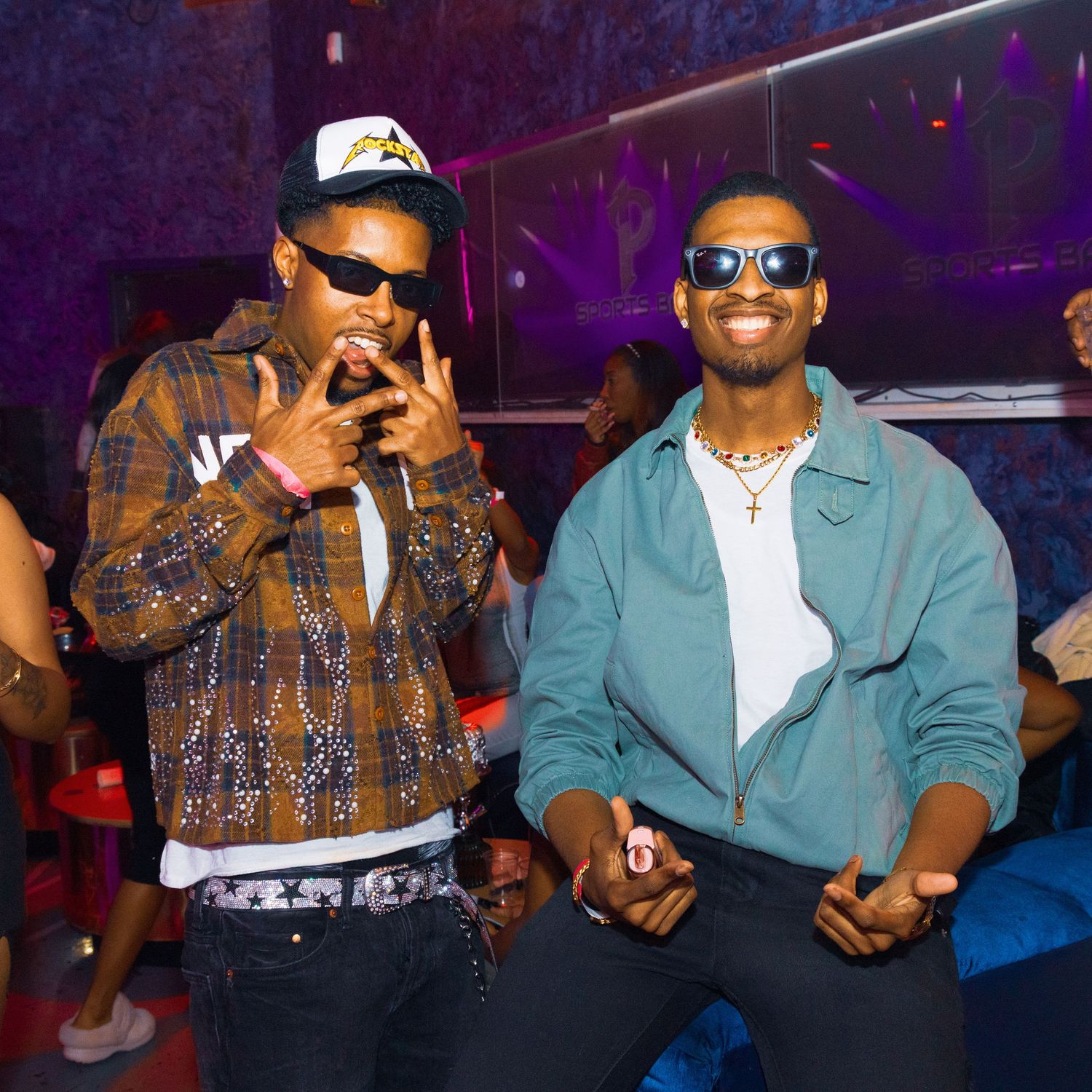 Two stylish young men posing together at a nightclub, one in plaid jacket and cap, other in teal jacket with gold chain.