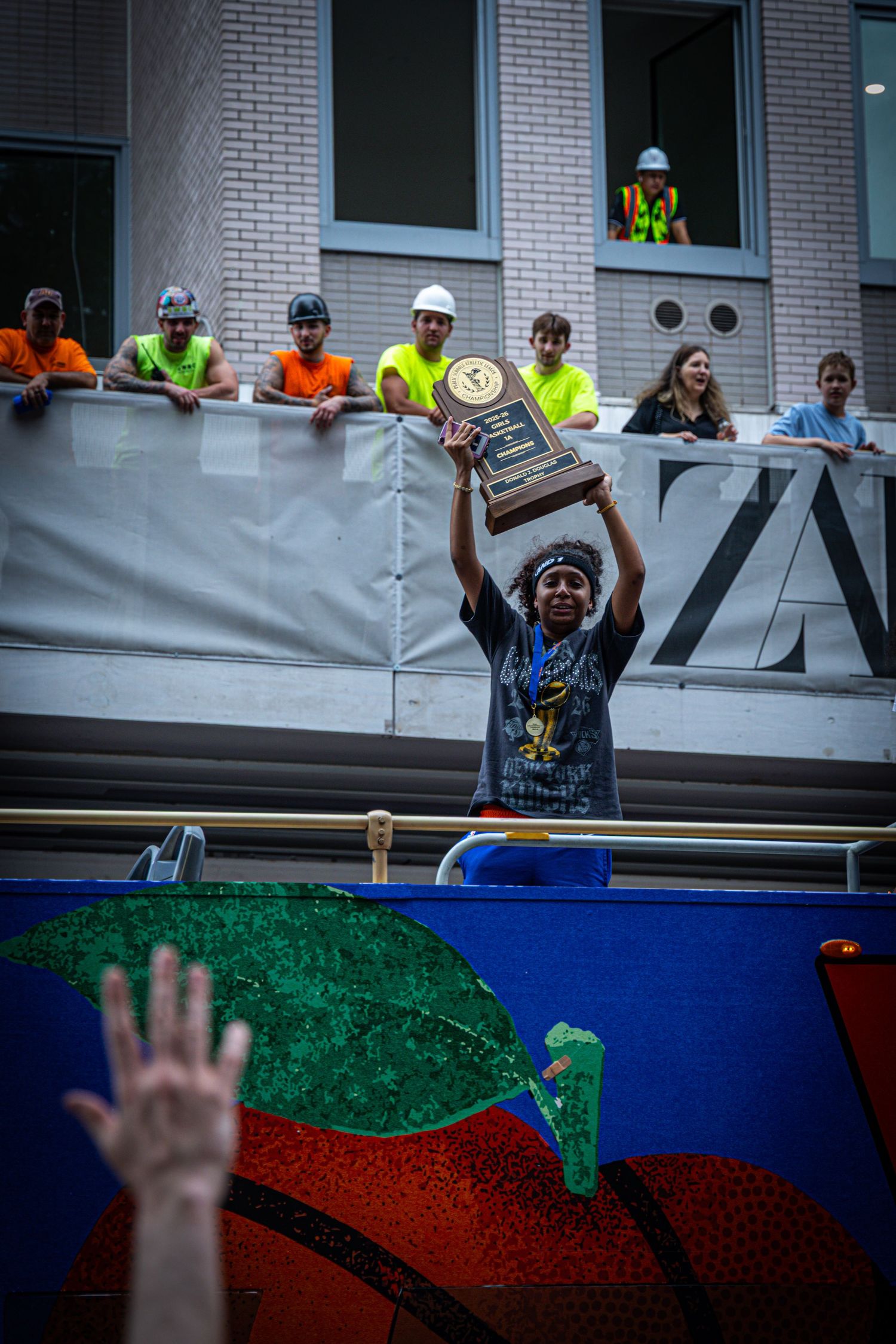 Athlete holds championship trophy aloft on parade float as construction workers watch from above.