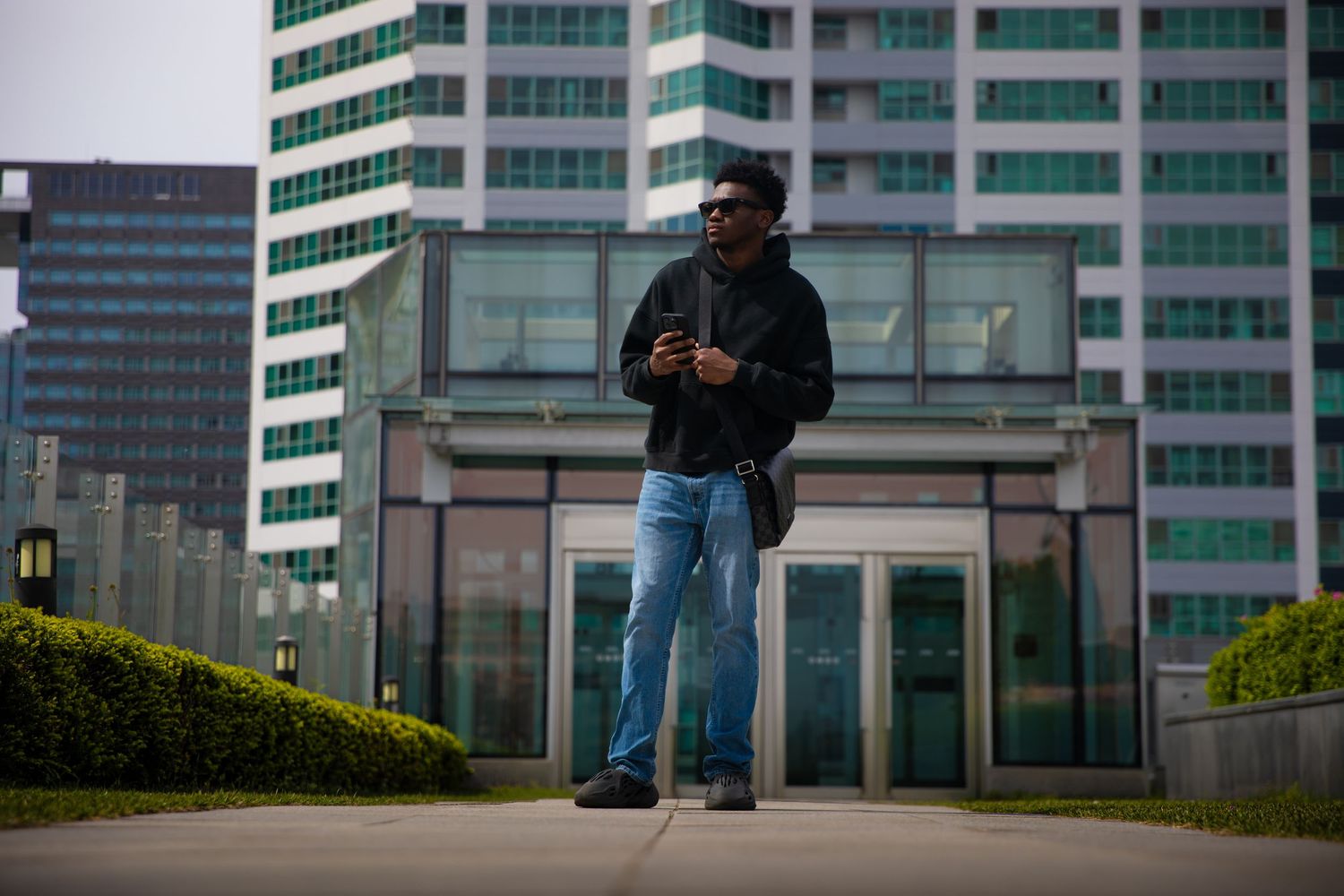 Man in black jacket and jeans wearing sunglasses standing outside modern glass office building holding smartphone.