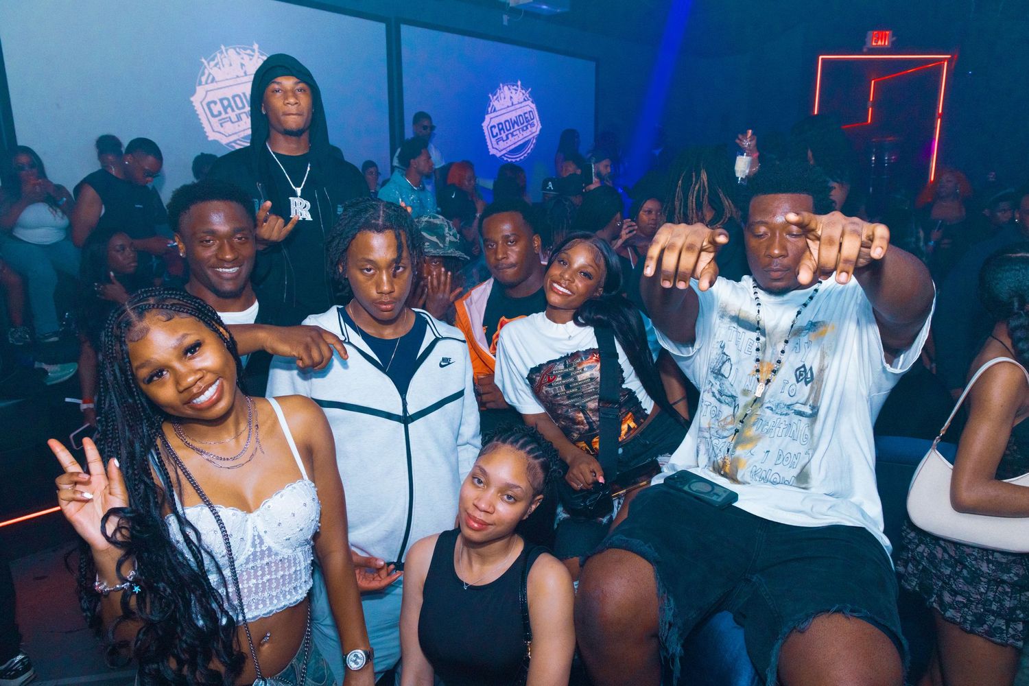 Group of young friends posing together at a nightclub party, smiling and making hand gestures under colorful lights.