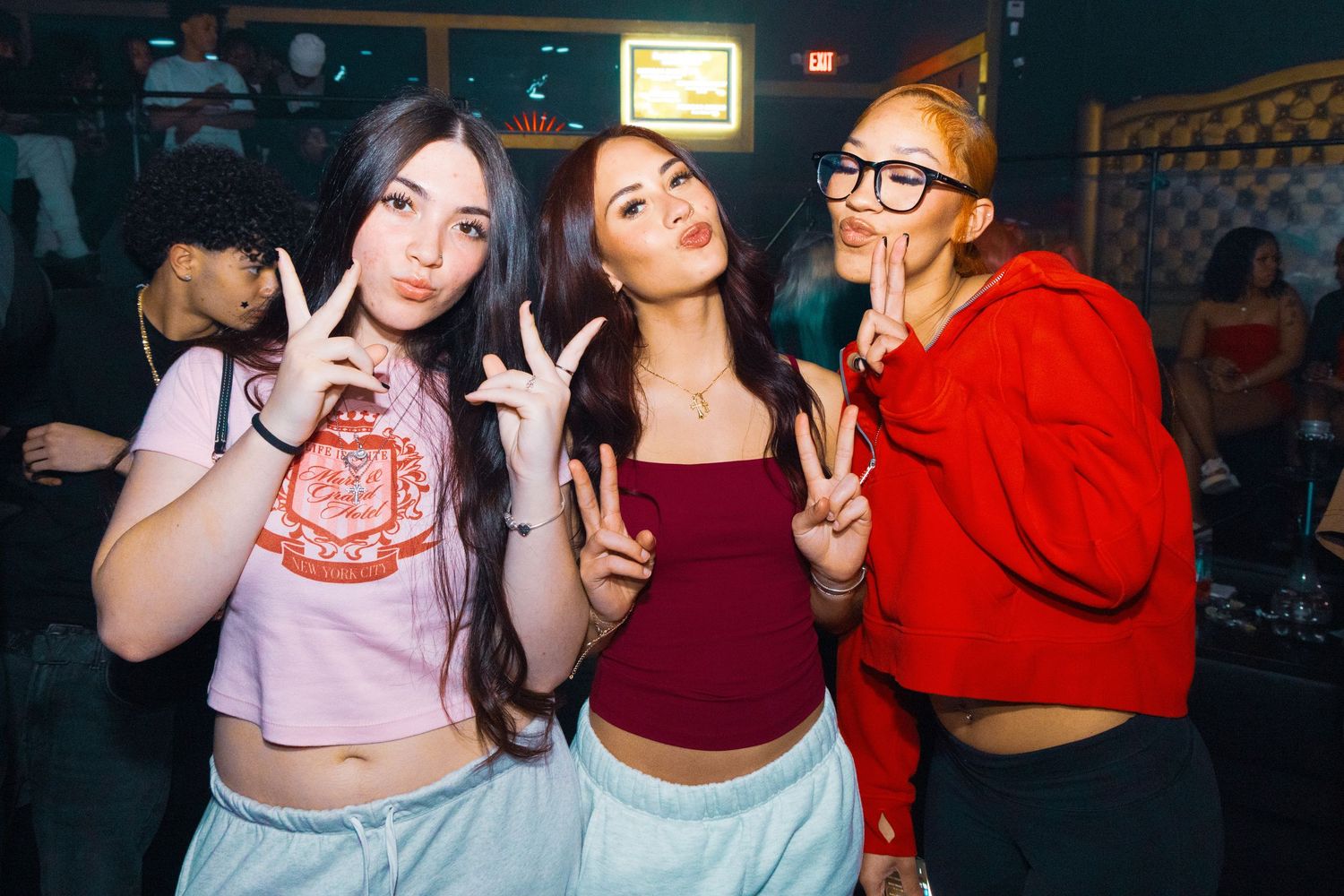 Three young women posing with peace signs at a nightclub, wearing casual outfits in a dimly lit venue.