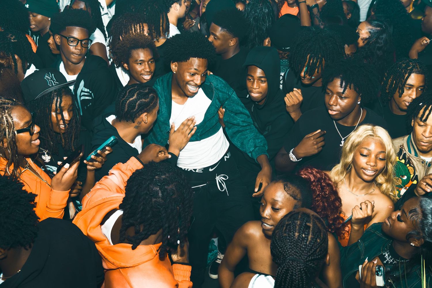 A young man in a green jacket is lifted by a crowd at a lively party with diverse guests.