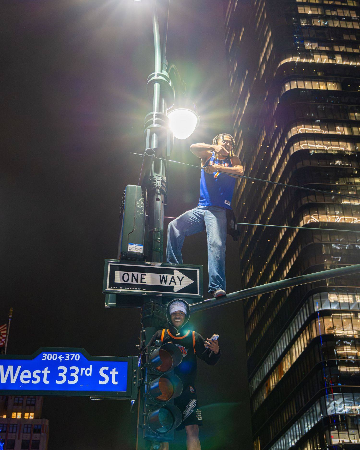 Two people climb a street pole at West 33rd St and One Way sign in New York City at night.