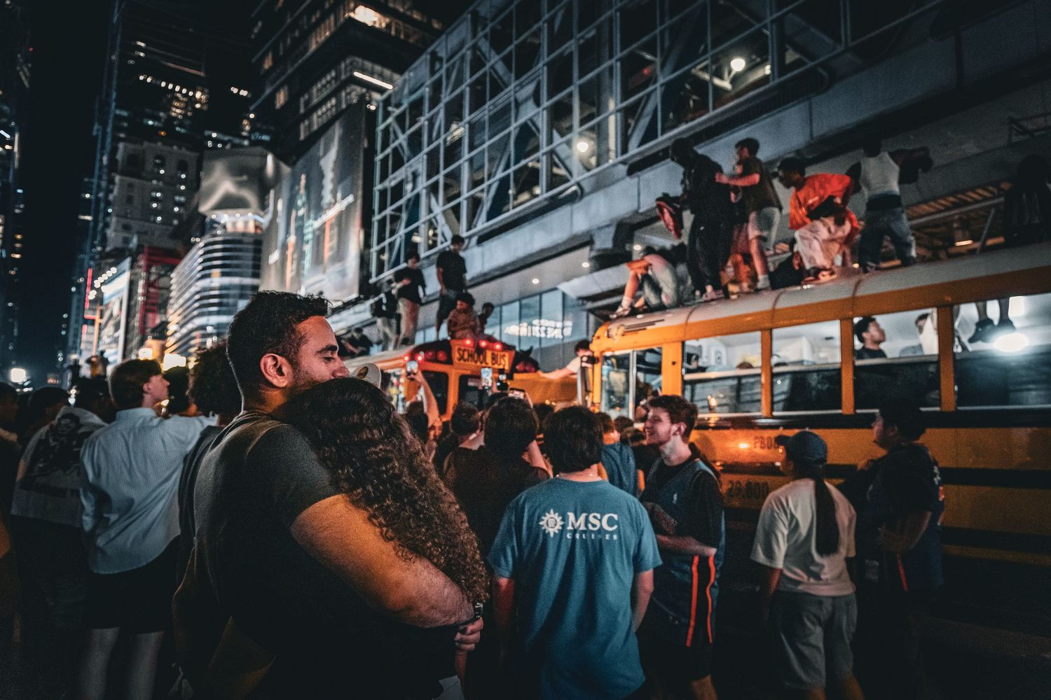 Crowd of people gathered at night around a yellow school bus on a busy city street with bright lights.