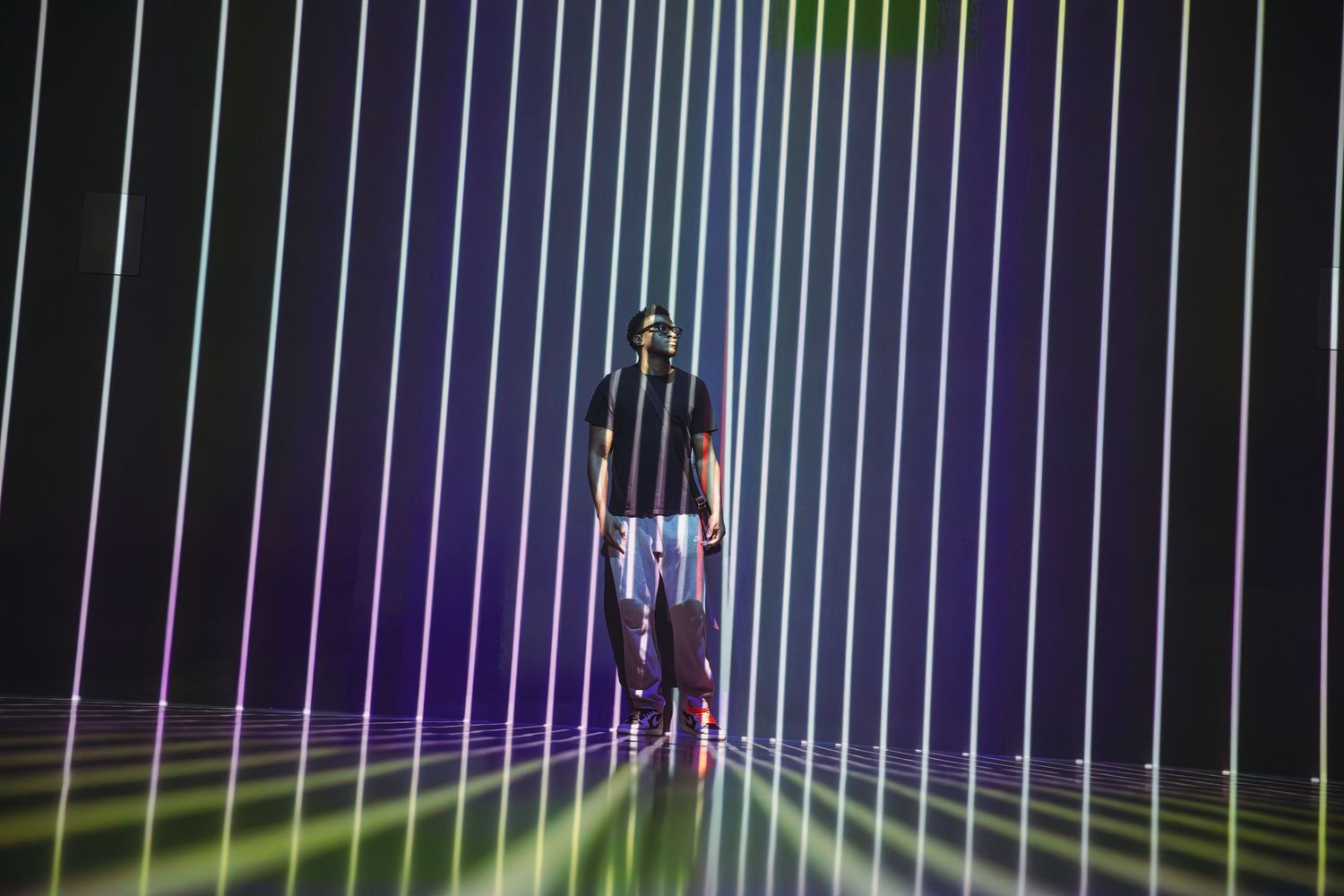 Person standing in colorful laser light show with vibrant green, purple, and blue beams on dark stage.