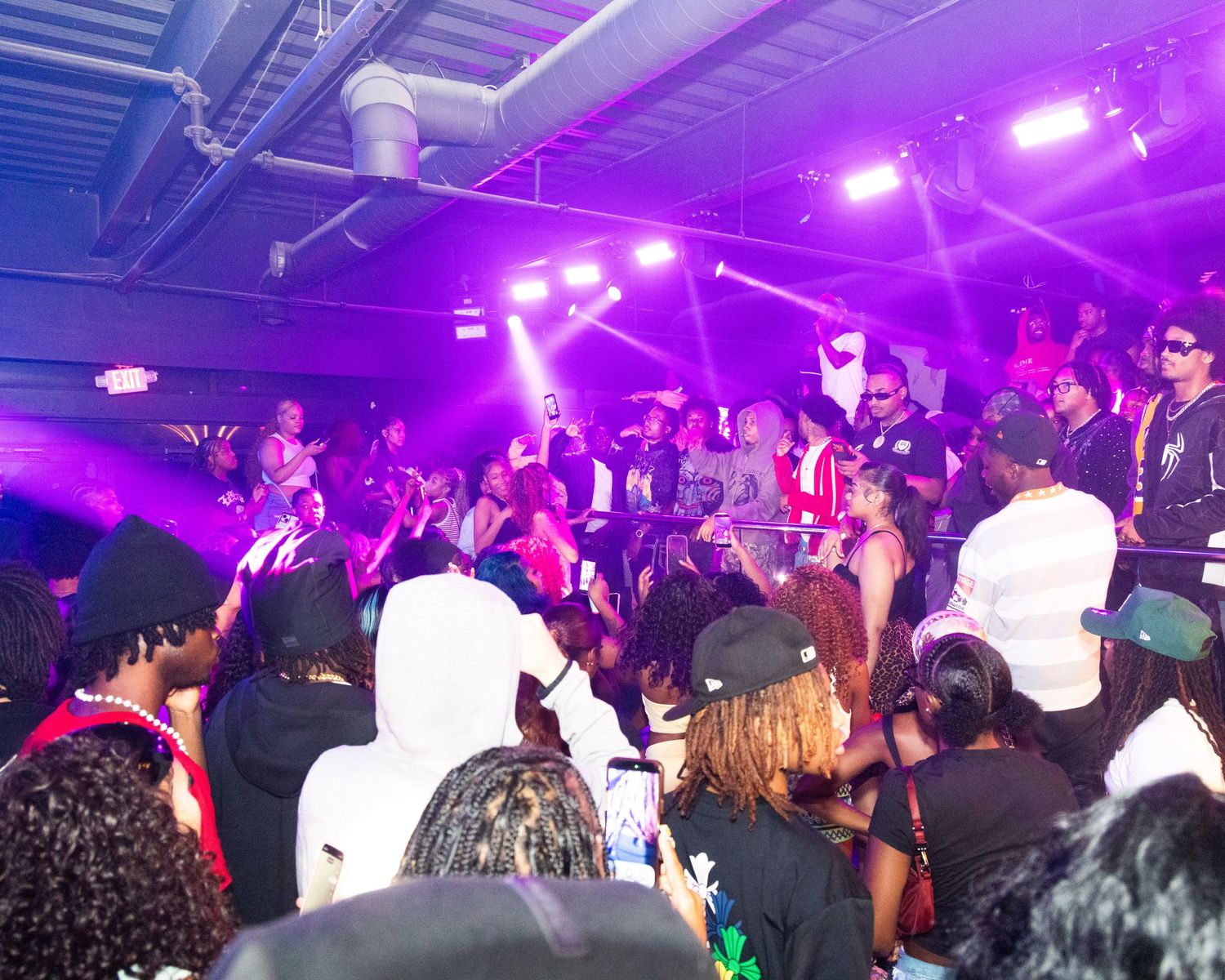 Crowded indoor party with purple stage lights, DJ, and diverse group of people dancing and socializing.
