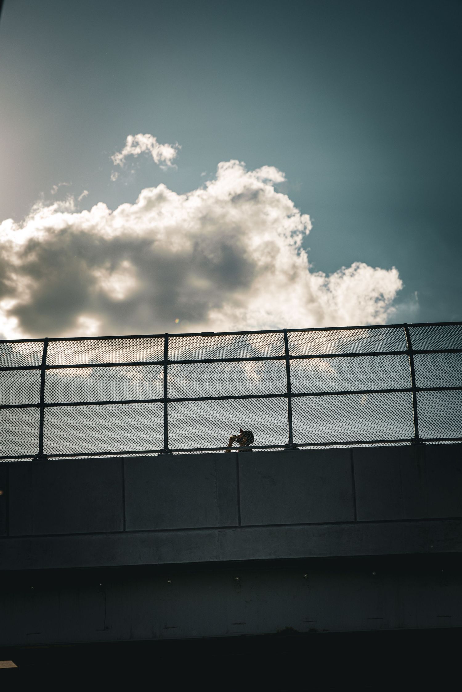 Silhouette of a person on a bridge railing against dramatic clouds and sunlight in a moody sky.