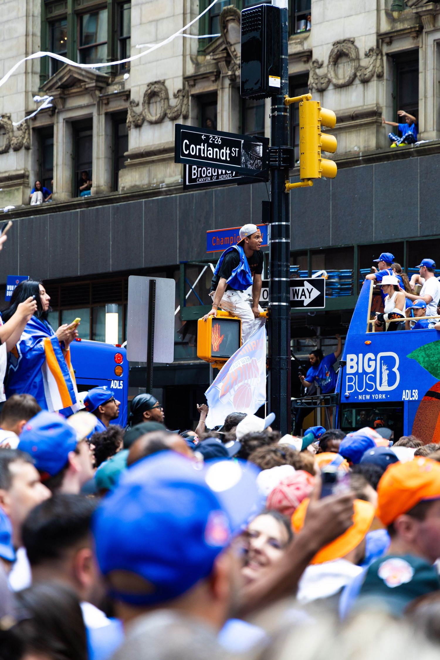 Crowd in blue hats at NYC parade on Broadway near Cortlandt St, with Big Bus and person on street sign.
