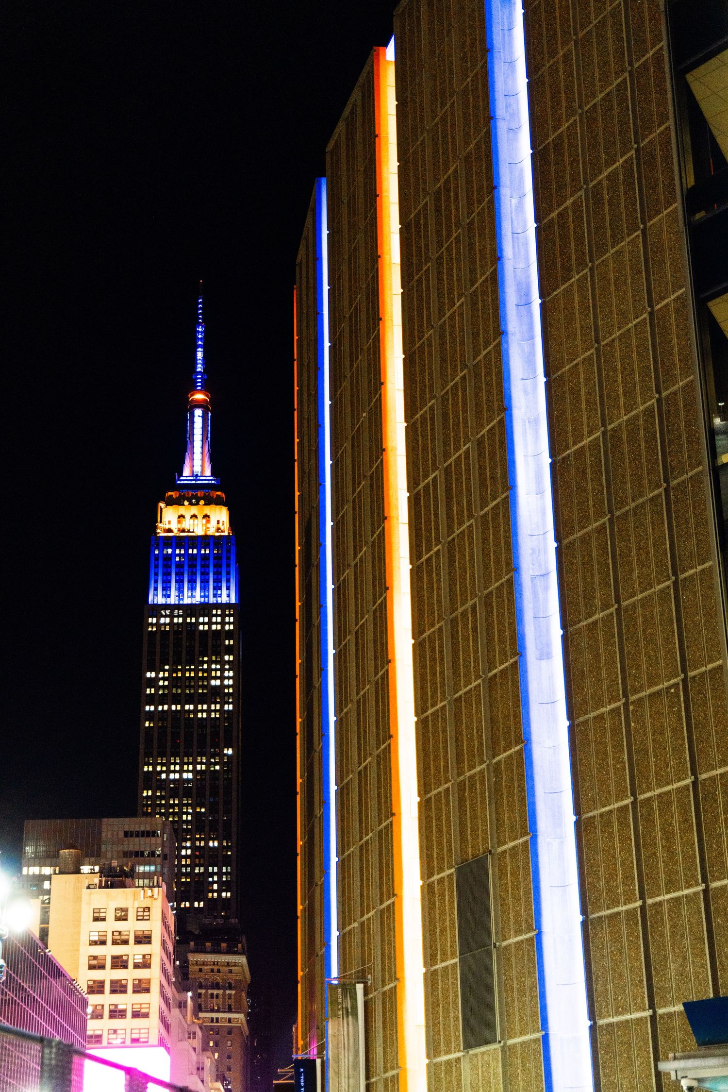 Empire State Building illuminated in blue and orange at night, flanked by modern buildings with neon light strips in NYC.
