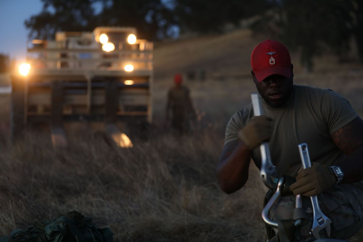 Soldier in red cap handling metal equipment in a field at dusk with military vehicle headlights in background.