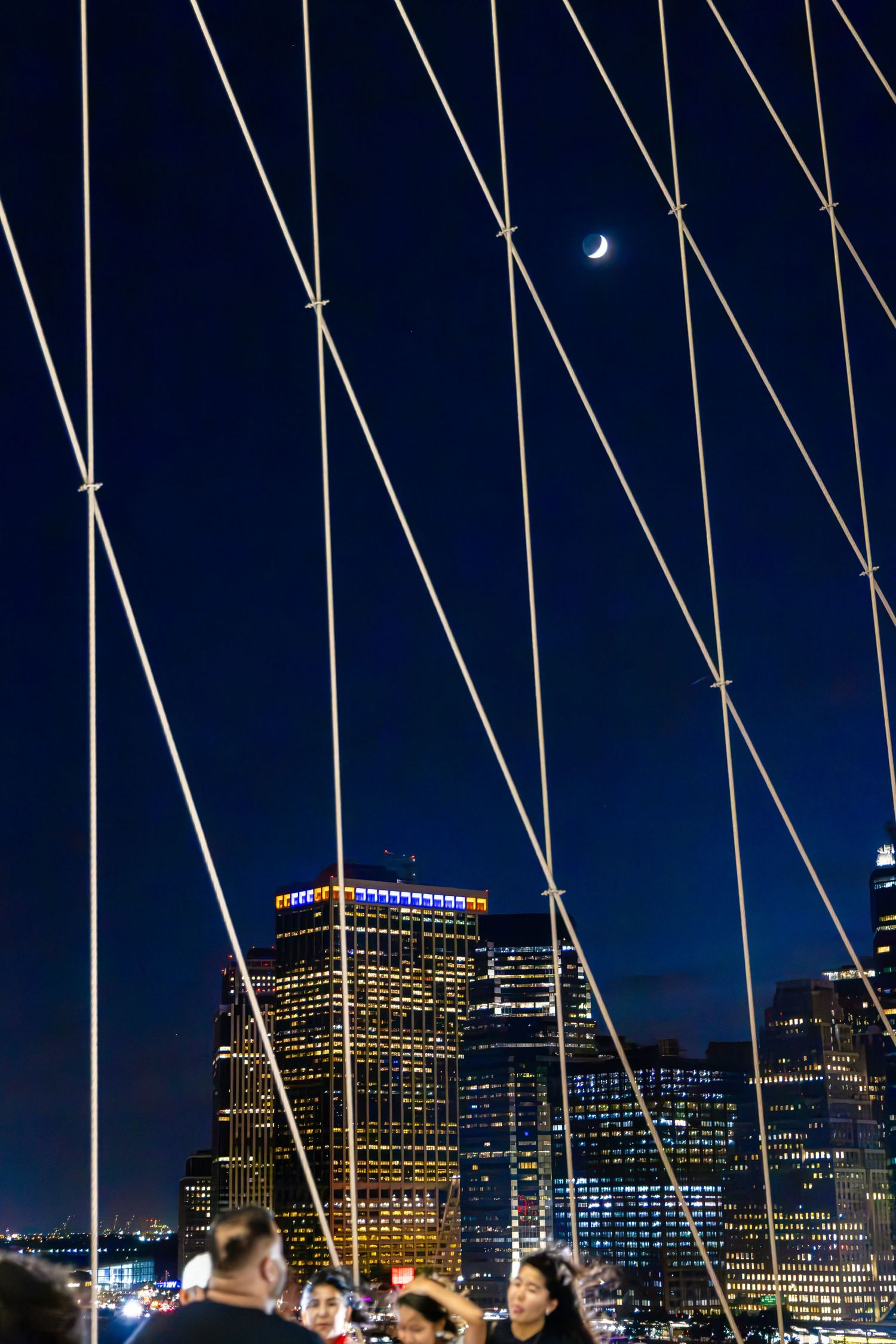 Night view through Brooklyn Bridge cables with crescent moon above illuminated Manhattan skyline and people gathered below.
