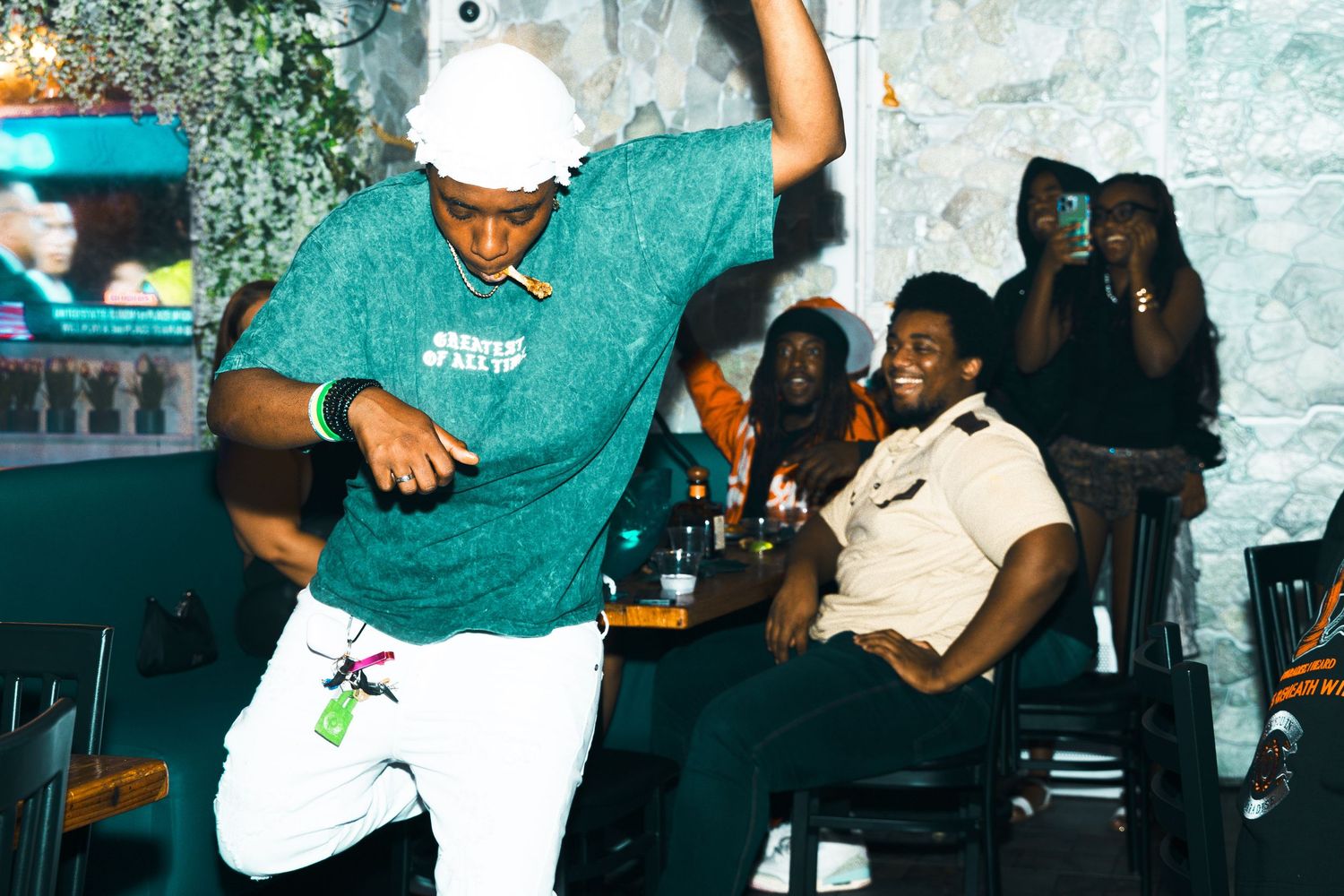 Man in green shirt dancing energetically at a lively bar while guests watch and smile in the background.