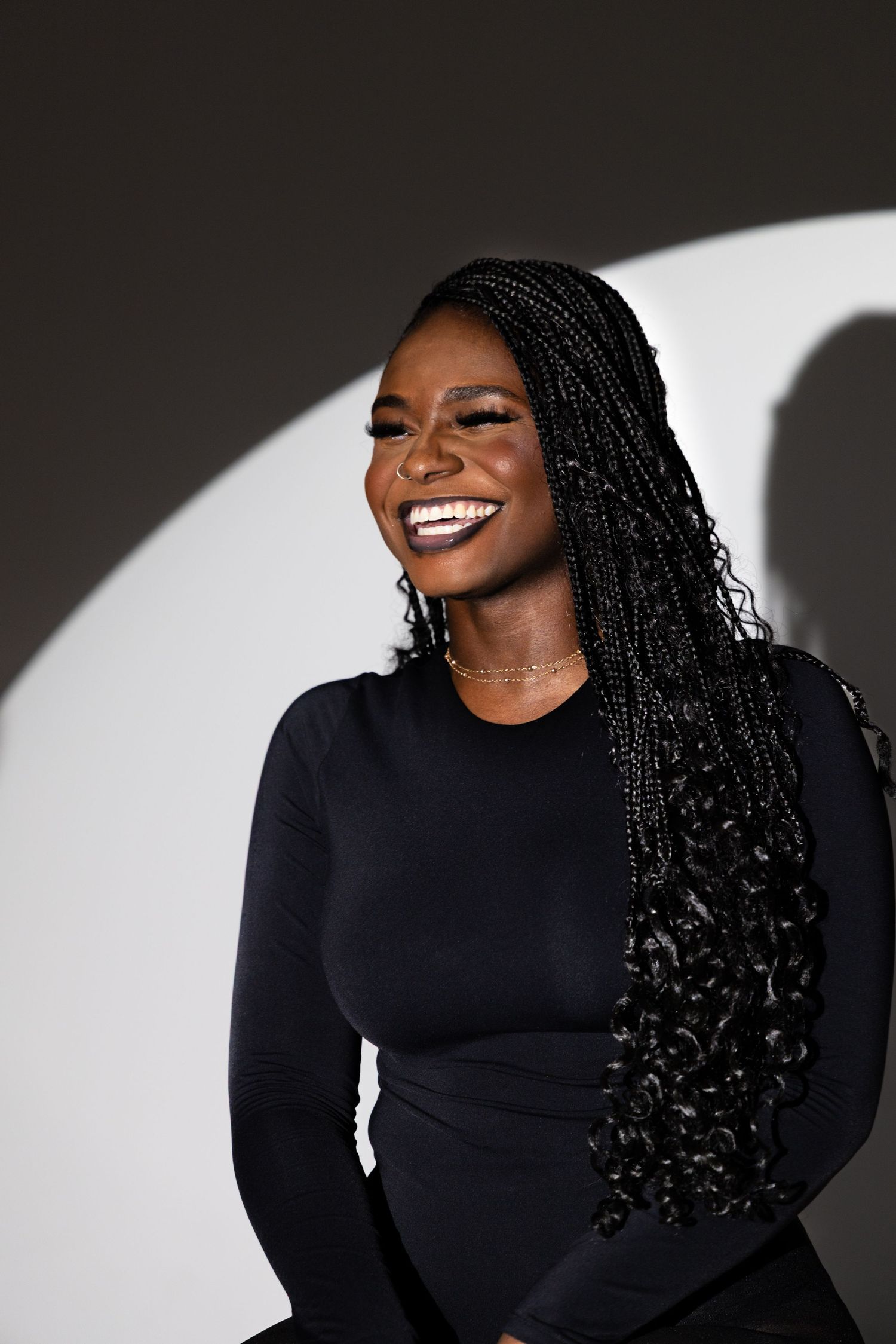 A smiling Black woman with long box braids wearing a black top, laughing against a grey studio background.