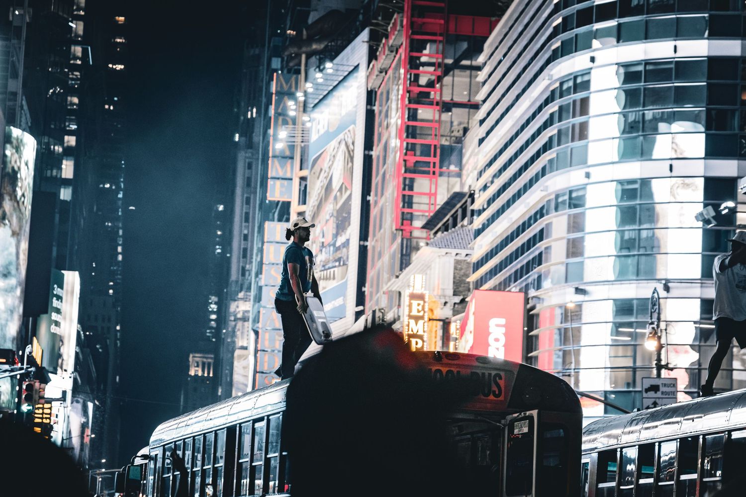 Person standing atop a dark structure at night in Times Square, NYC, surrounded by glowing billboards.