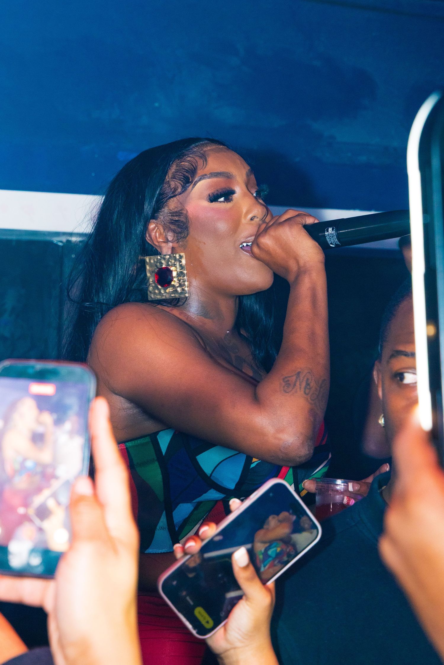 Female performer singing into microphone on stage, surrounded by fans recording with smartphones at a nightclub event.