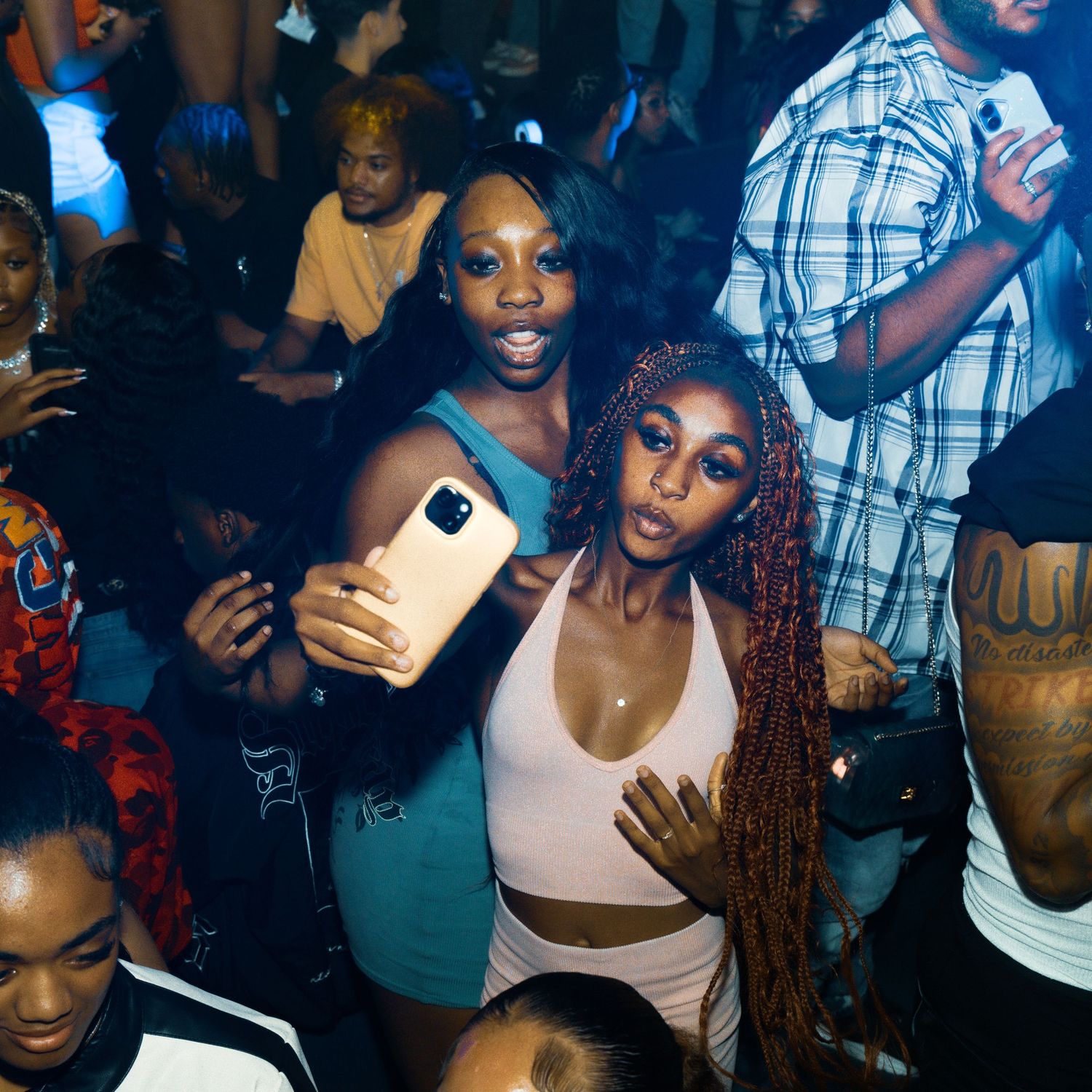 Two women pose for a selfie at a crowded nightclub party, surrounded by other guests in a dimly lit venue.