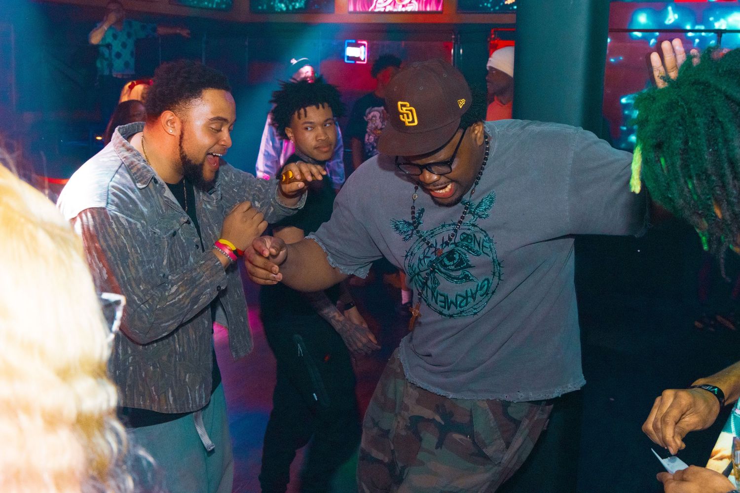 Two men dancing and laughing together at a lively nightclub with colorful neon lights.