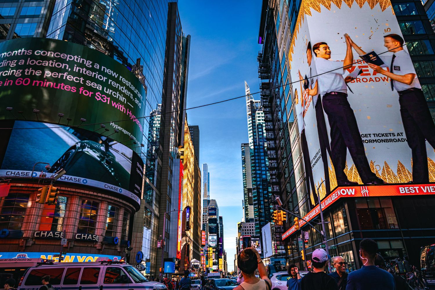 Busy Times Square NYC at dusk with illuminated billboards, skyscrapers, and crowds of people on the street.